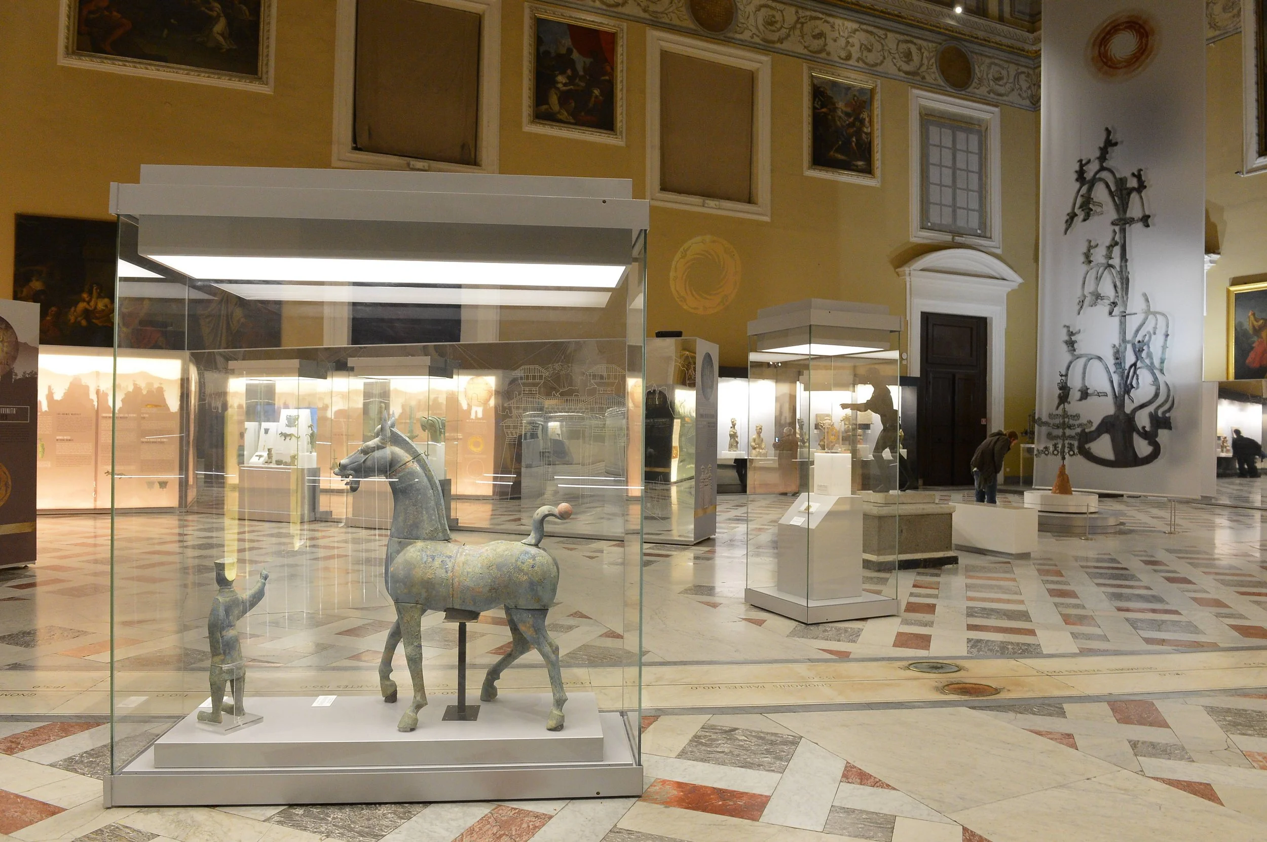 An art museum interior with glass display cases containing ancient sculptures, including a horse figure and human figures. The room features artistic wall paintings and a large wall mural with black ink illustrations. The floor is marble with a geome