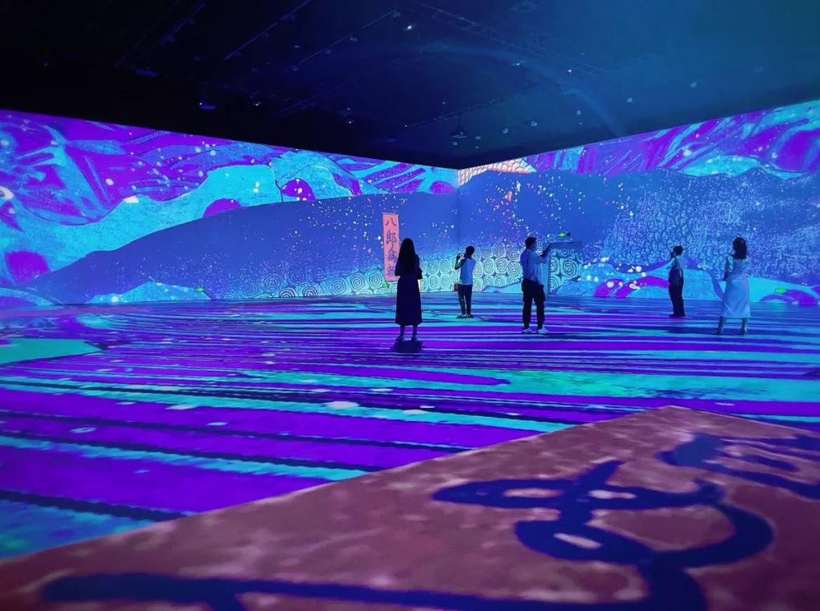 People standing and interacting inside an immersive digital art installation with blue and purple light projections on the walls and floor.
