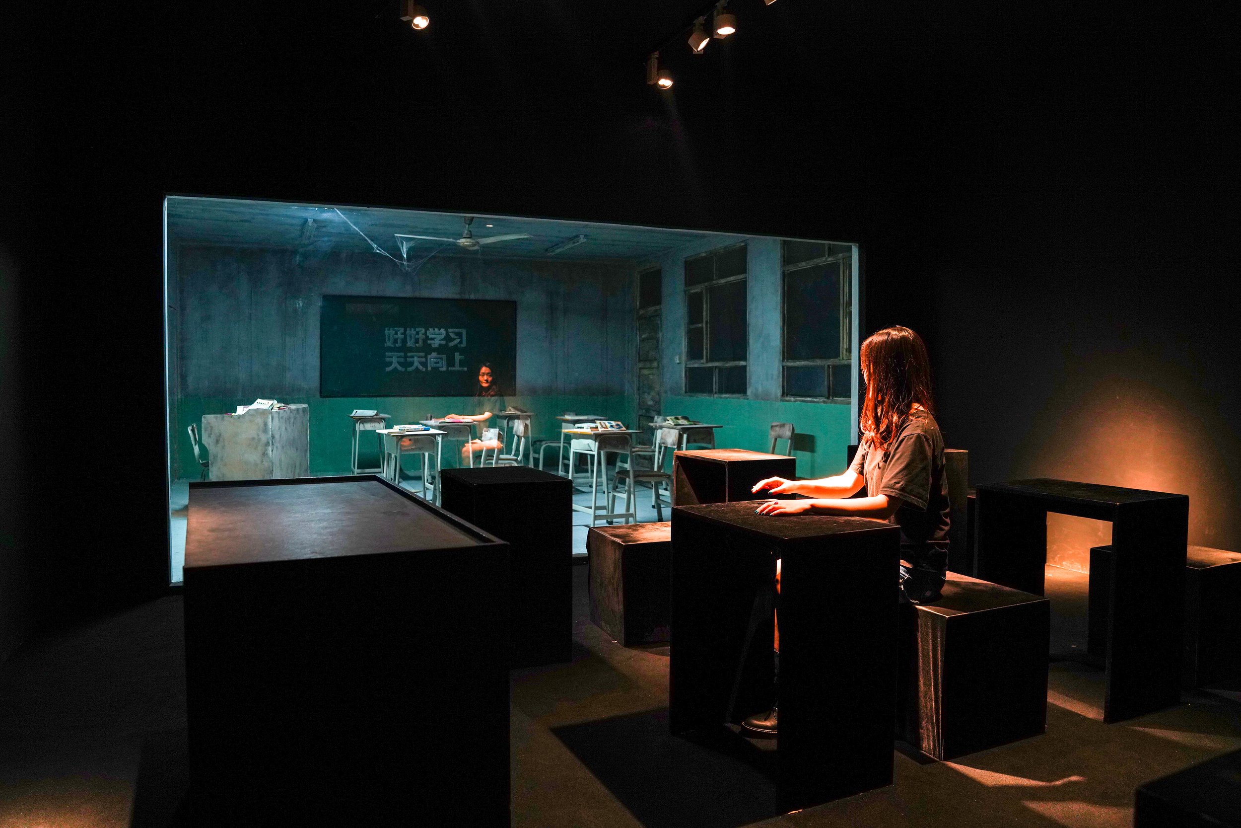 A woman sitting on a black cube in a dark room, looking at a large window into an empty classroom with desks and chairs, and a person sitting at a desk on the other side.