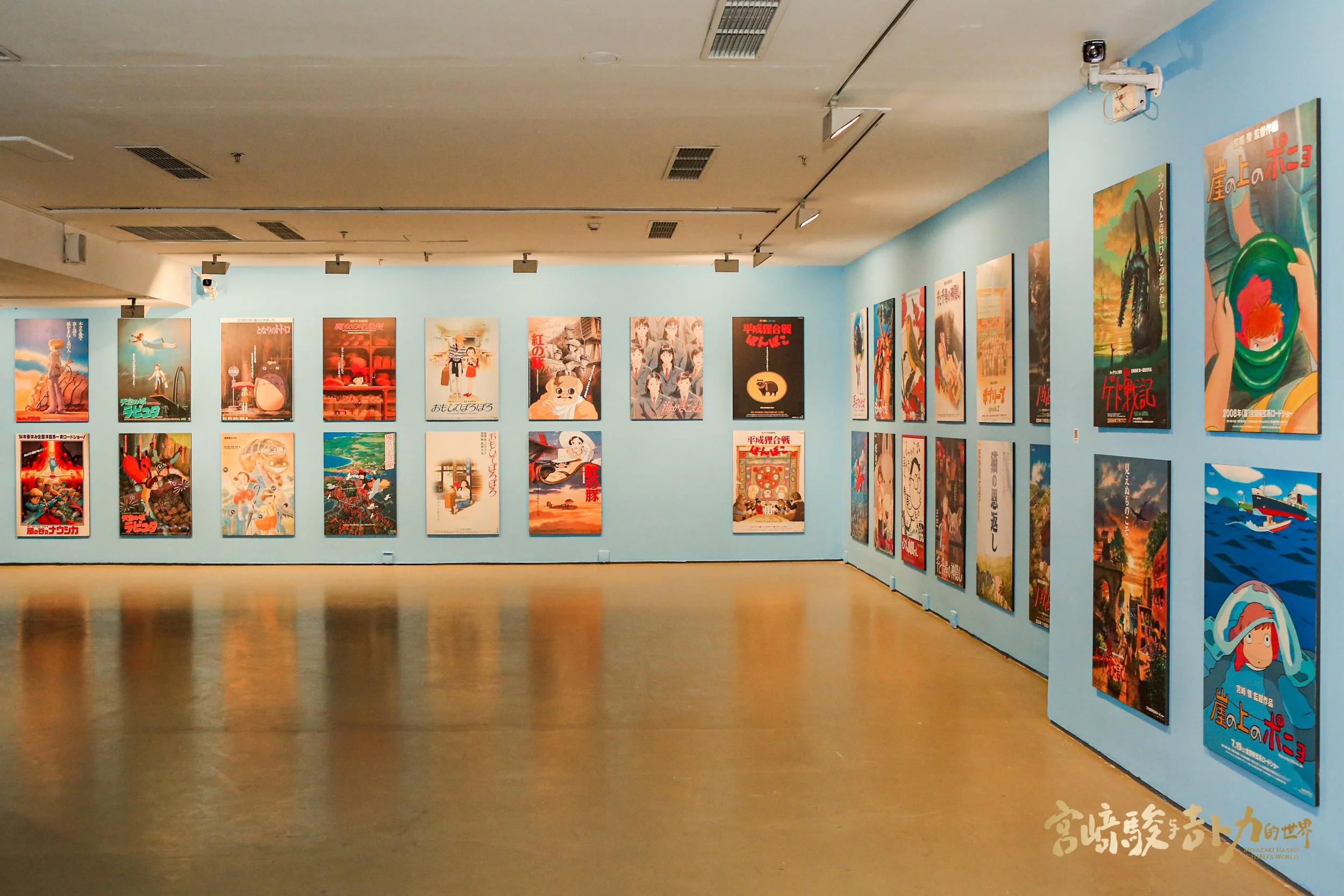 An art gallery with blue walls displaying multiple posters of animated films, arranged in rows and columns, with track lighting overhead.