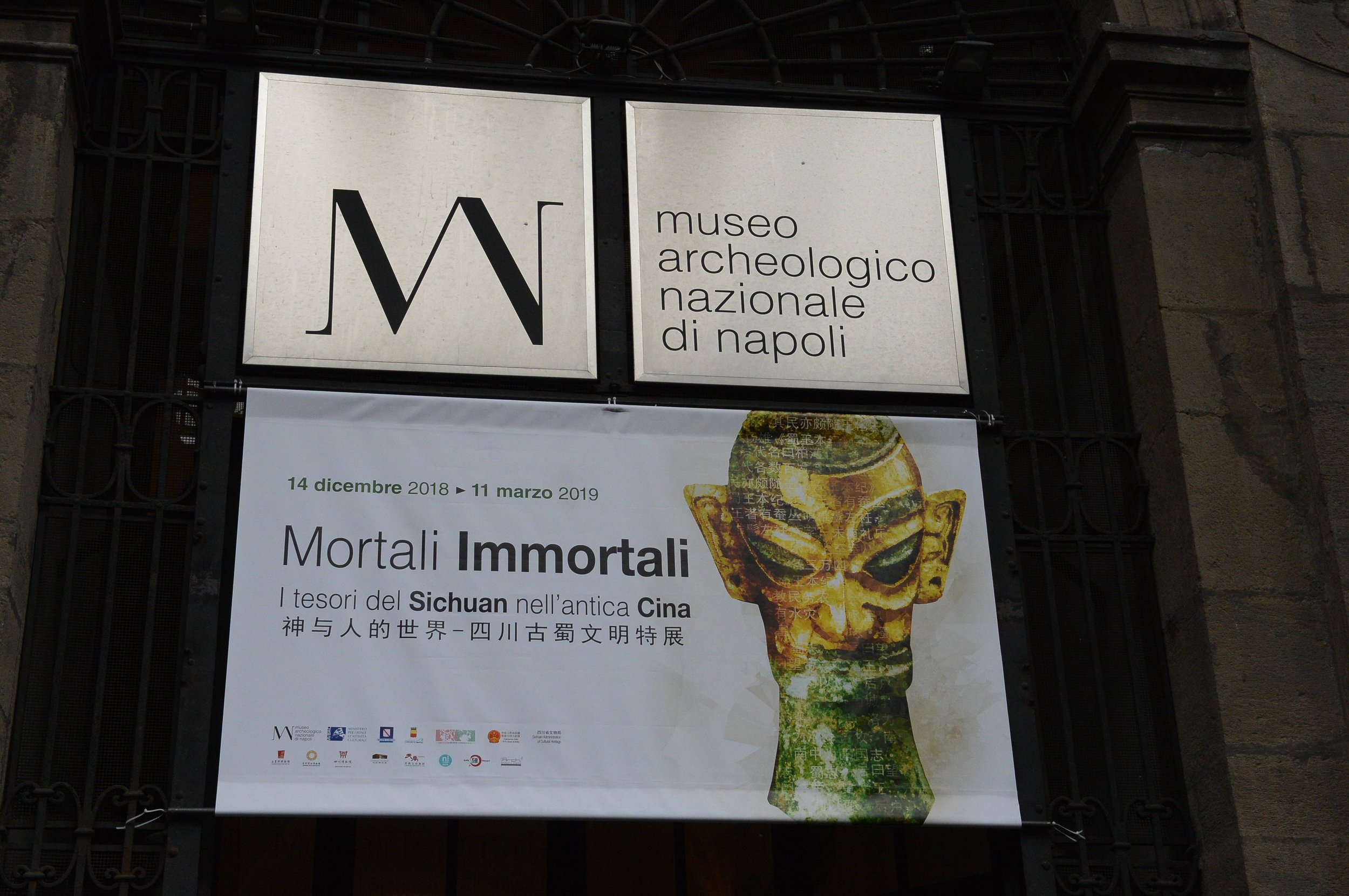 Signboard for the Museo Archeologico Nazionale di Napoli with an exhibition poster about Mortali Immortali, displaying a stylized ancient mask and text in Italian, English, and Chinese.
