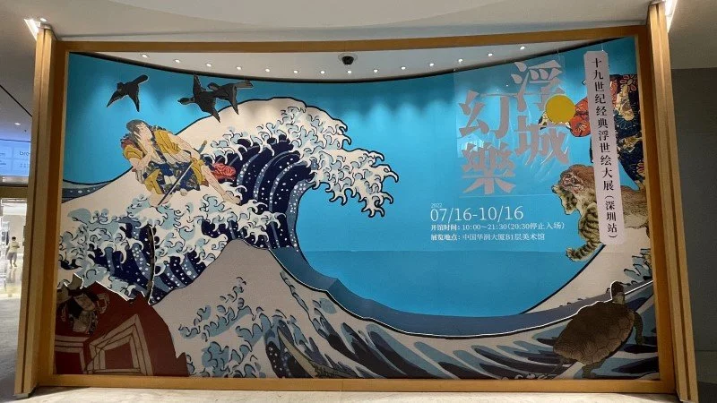 Poster featuring the famous Japanese artwork The Great Wave off Kanagawa, with modern elements like sharks, a plane, and a turtle, combined with text in Chinese and Japanese promoting an exhibit on ocean-themed art from July 16 to October 16.