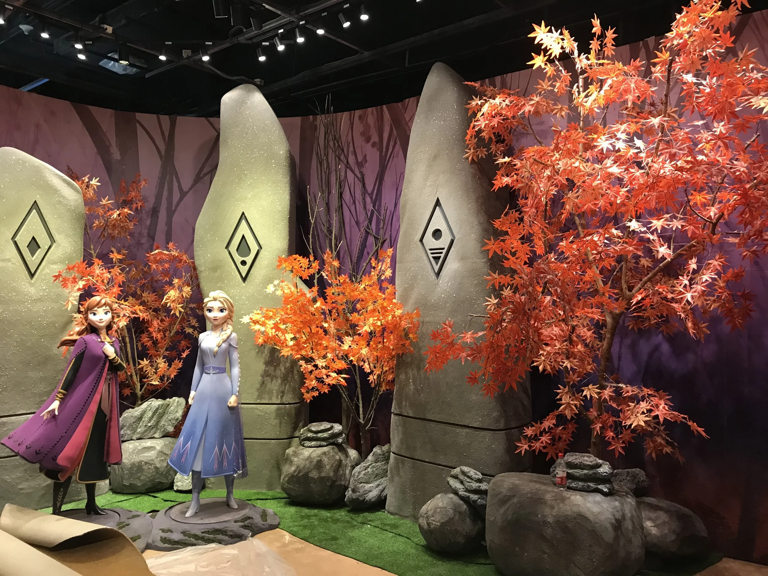 Disney's Frozen themed scene with orange autumn trees, large rocks, and statues of Anna and Elsa in front.