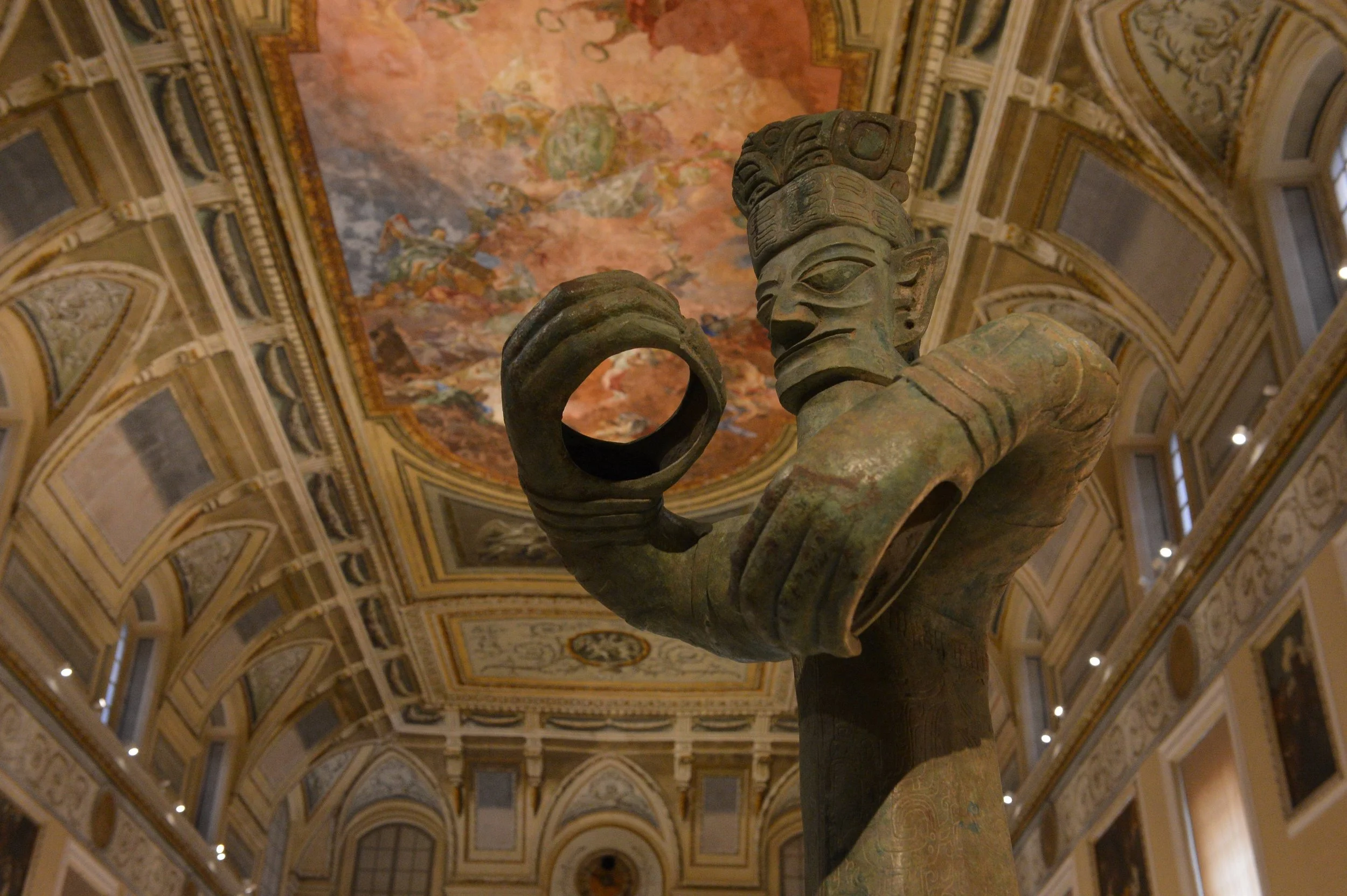 Ancient sculpture of a humanoid figure with an elaborate headdress, holding a circular object in one hand, located in a ornate museum hall with detailed ceiling artwork.