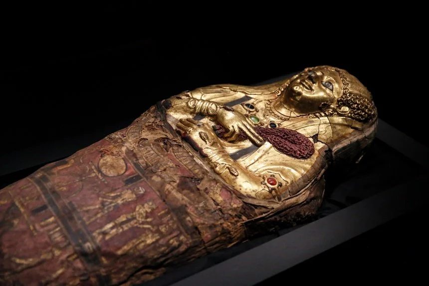 Ancient Egyptian coffin with a painted and gilded portrait of a woman, decorated with jewelry and hieroglyphics, displayed in a museum setting.
