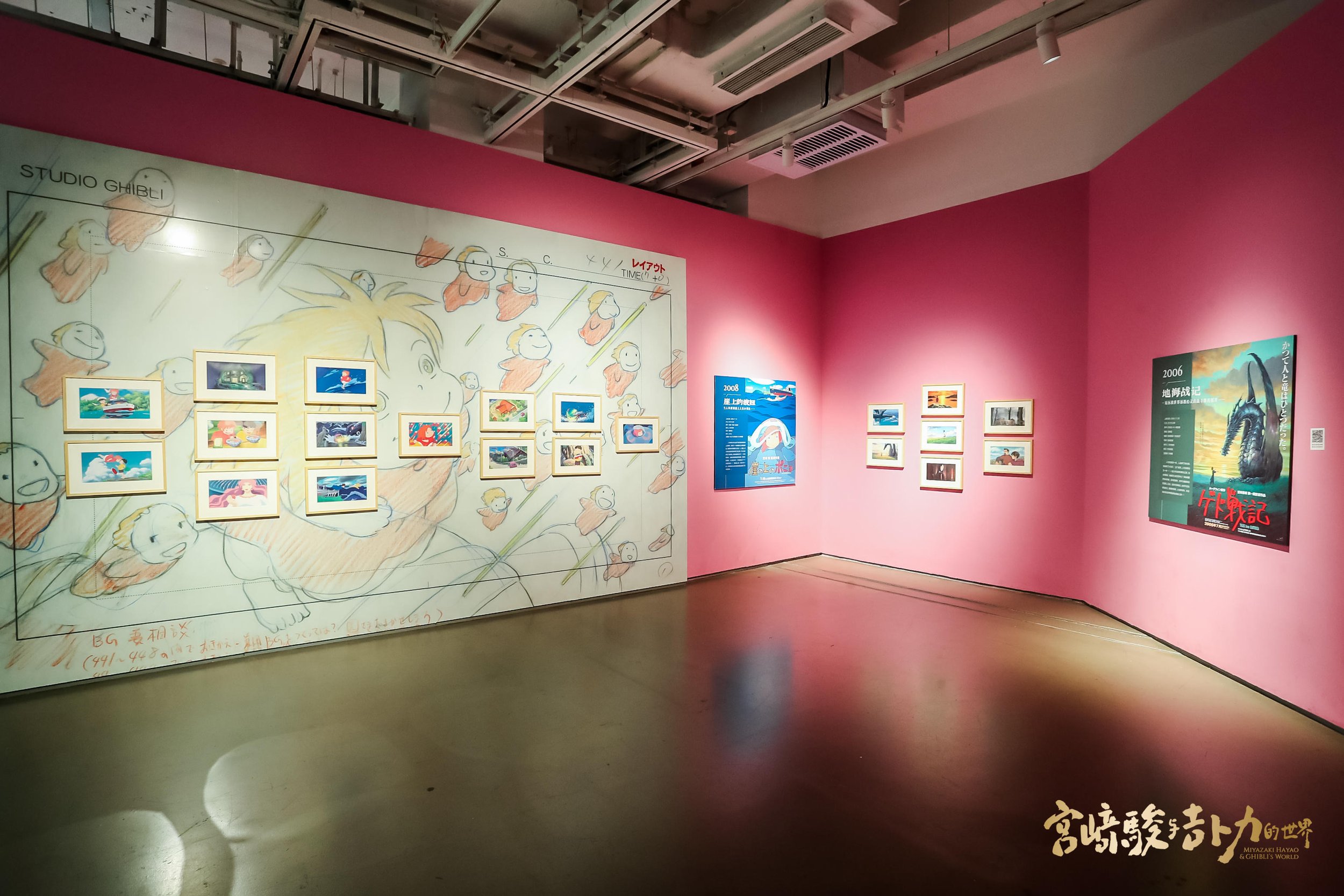 An art gallery with pink walls displaying framed animated film posters and artwork, including a large background sketch of a character and several smaller framed pictures, some of which are from Studio Ghibli films.