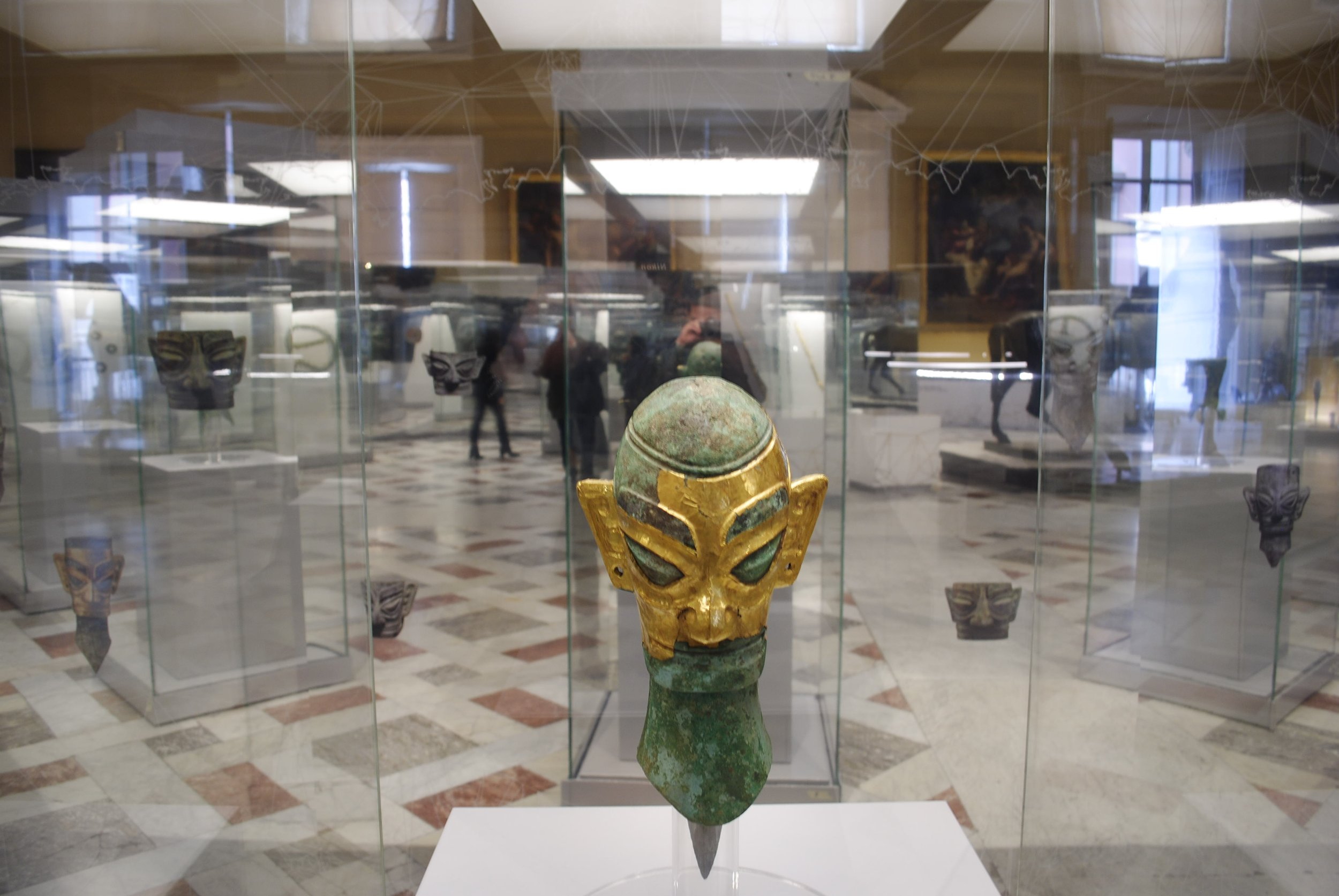 Ancient metallic mask with golden face and greenish head, displayed in a glass case at a museum.