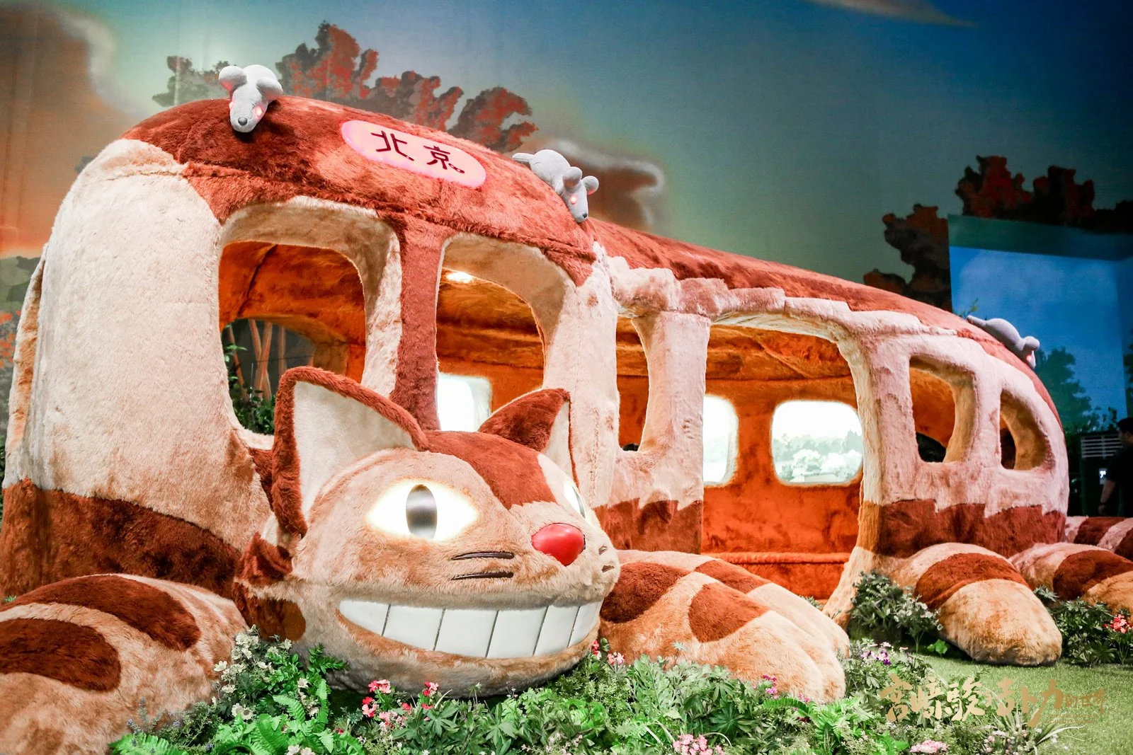 A large, plush, cartoonish bus and cat sculpture, both with smiling faces and vibrant colors, are displayed indoors with decorative plants and flowers around them.