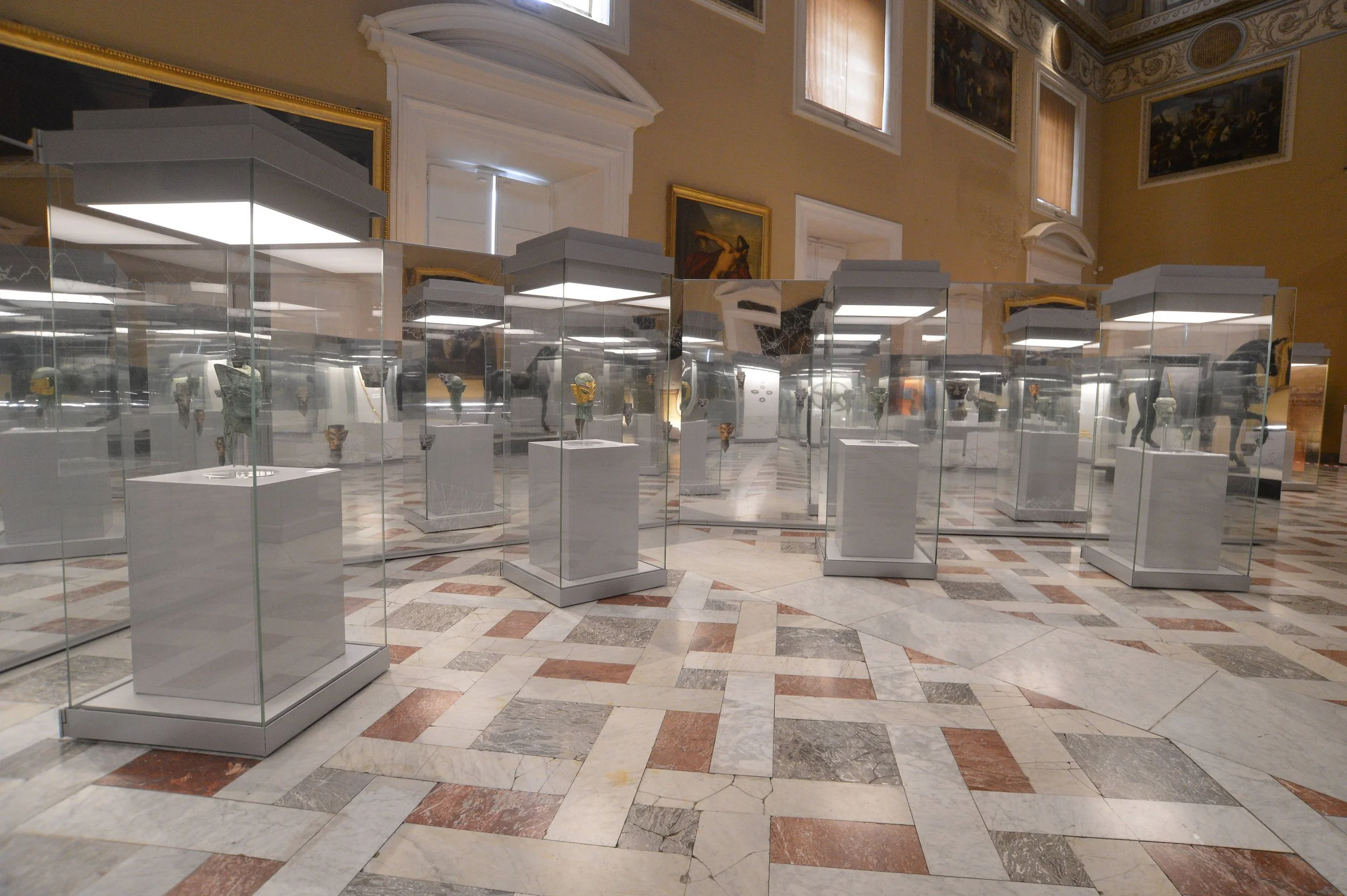 Museum exhibit with glass display cases on pedestals holding ancient artifacts in a historic gallery with high ceilings, paintings, and intricate wall decorations.