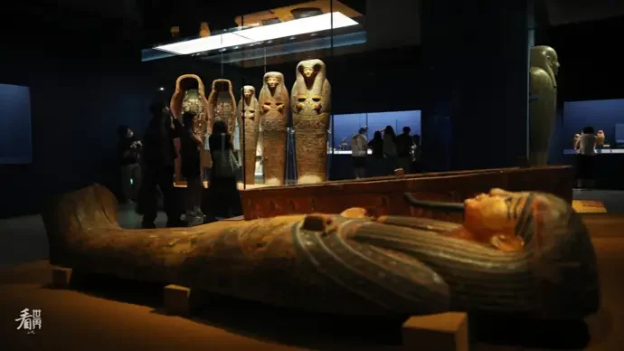 Ancient Egyptian artifacts, including a wooden sarcophagus and several painted mummies in a museum exhibit.