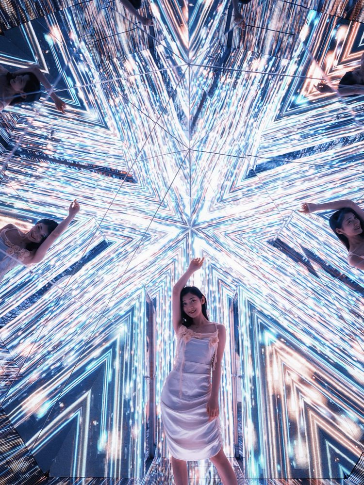 Woman in white dress posing inside a mirrored room with bright, colorful, star-shaped LED lights reflecting off the walls creating a symmetrical, kaleidoscopic pattern.