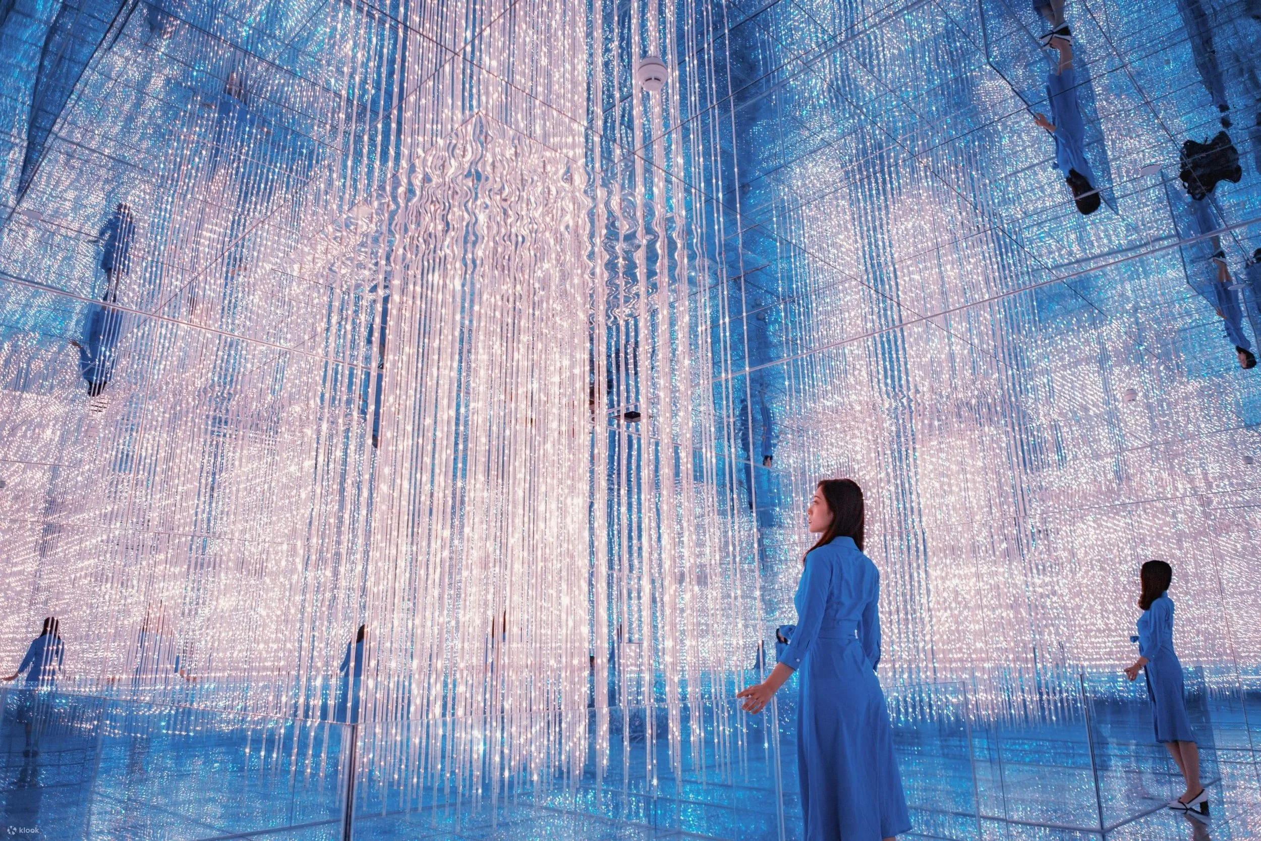 A woman in a blue dress standing inside an immersive mirror installation with hanging LED lights creating a shimmering, multi-directional light display.