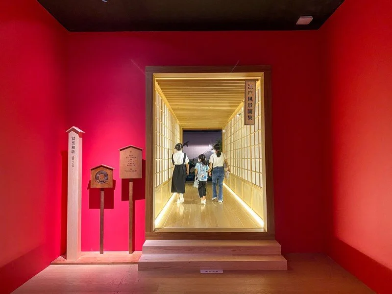 A group of four children and one adult walking through a traditional Japanese wooden corridor entrance with shoji sliding doors, inside a room with bright pink walls.