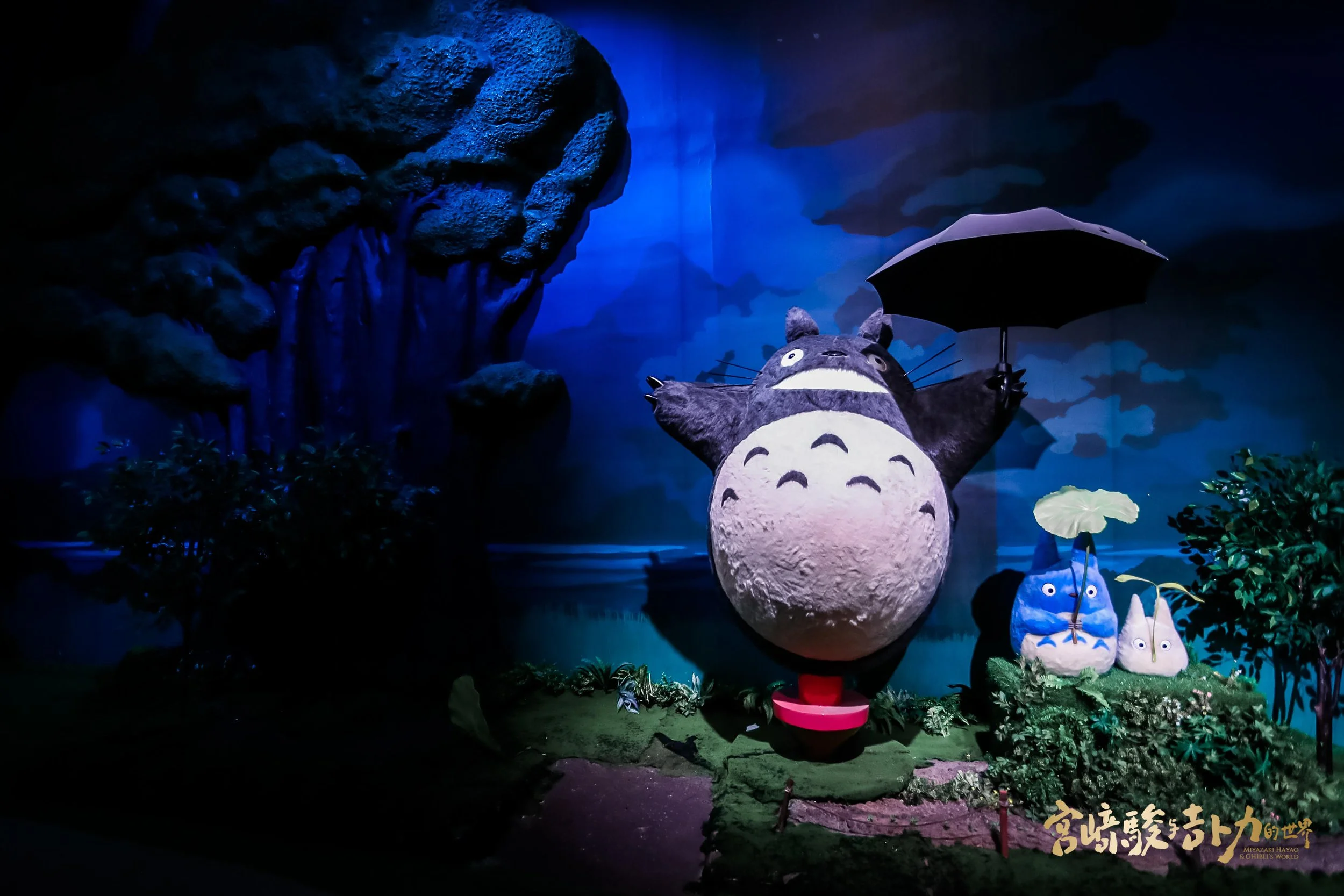 A large Totoro character holding a black umbrella, surrounded by small Totoro figures holding a yellow umbrella, set against a dark, mystical forest background.