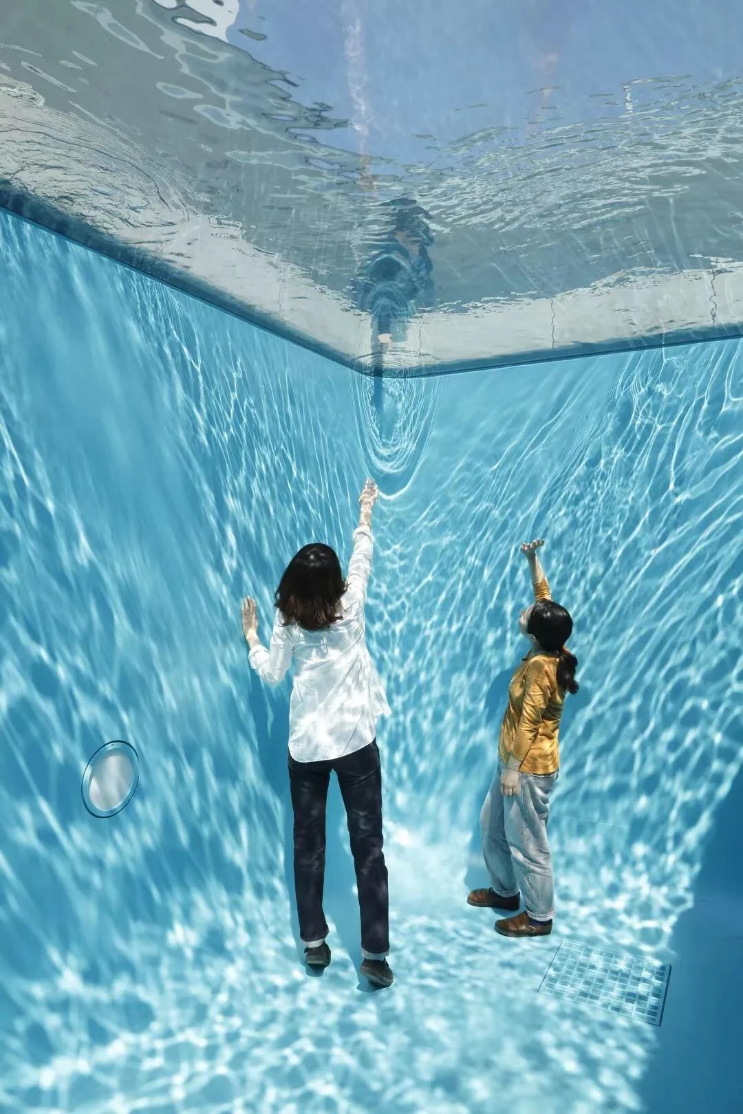 Two women are underwater in a shallow swimming pool, reaching out towards the ceiling with one arm each.