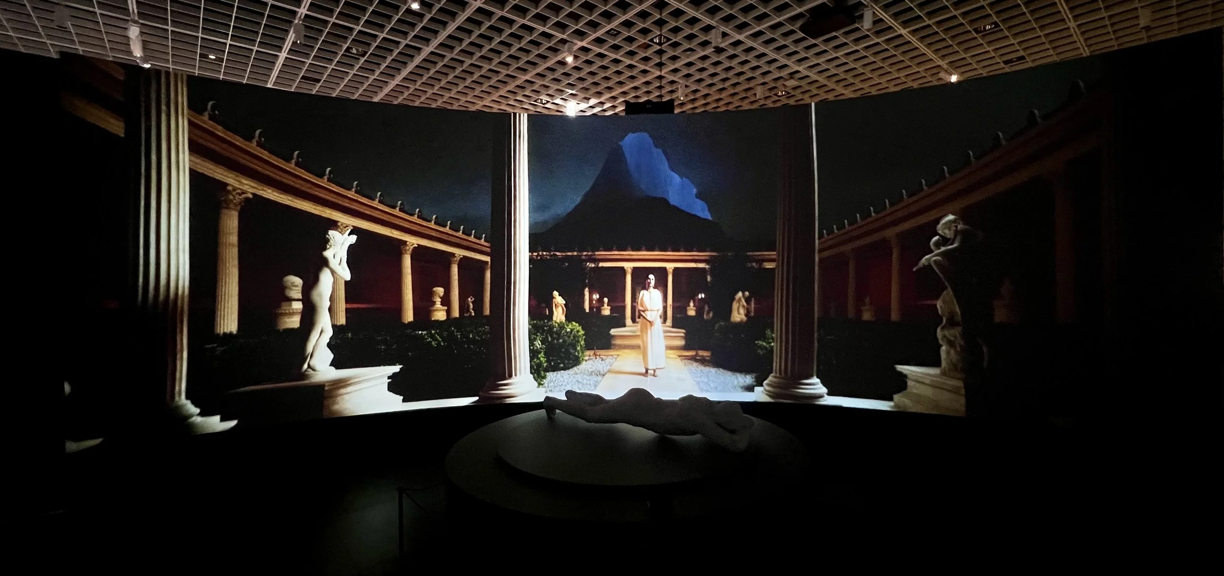 A dark art installation resembling an outdoor classical Greek garden with statues and columns, under a night sky with a mountain in the background, and a woman in a white dress standing in the center.