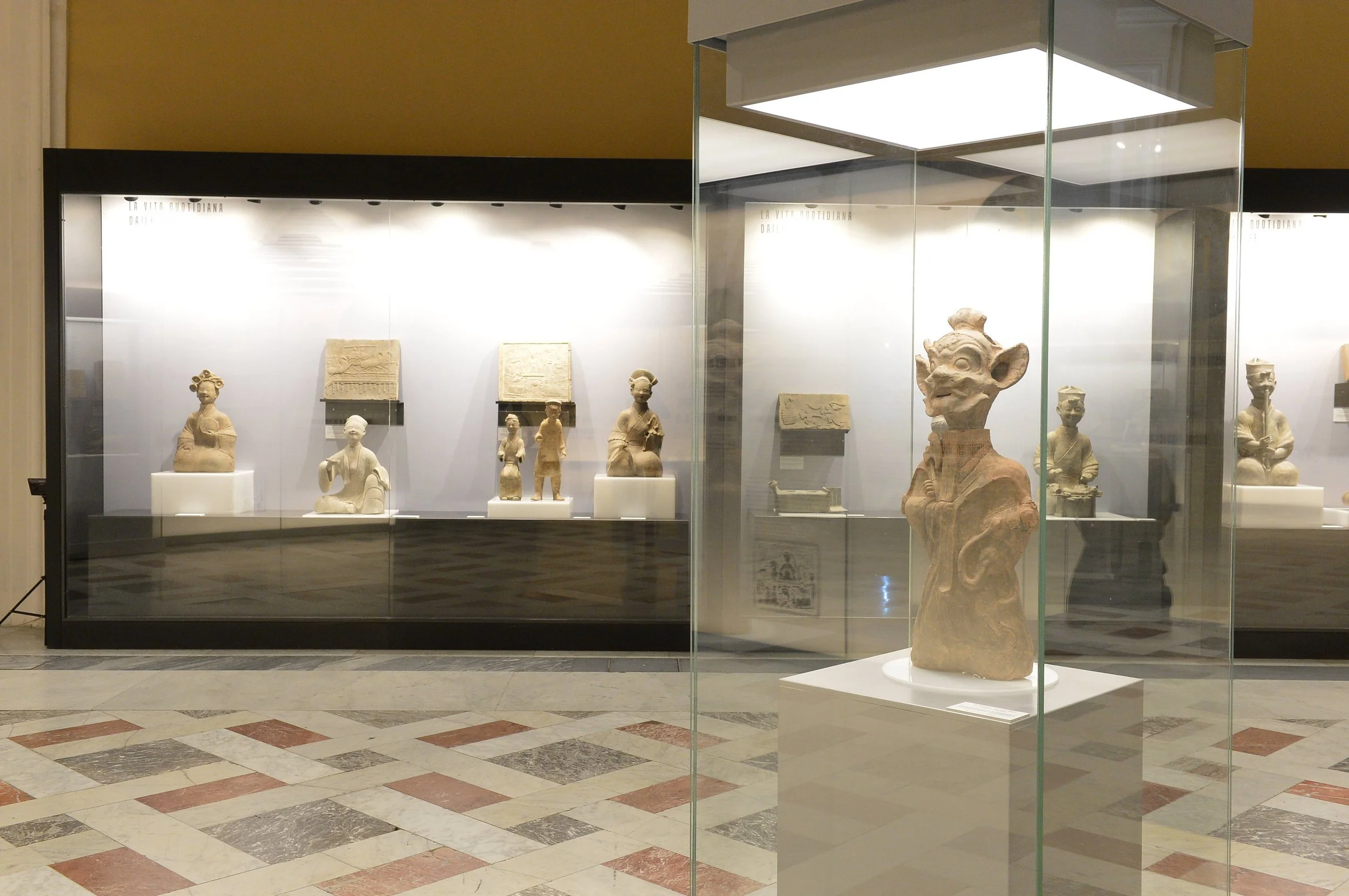 Museum display with ancient sculptures in glass cases against a beige wall, marble floor.