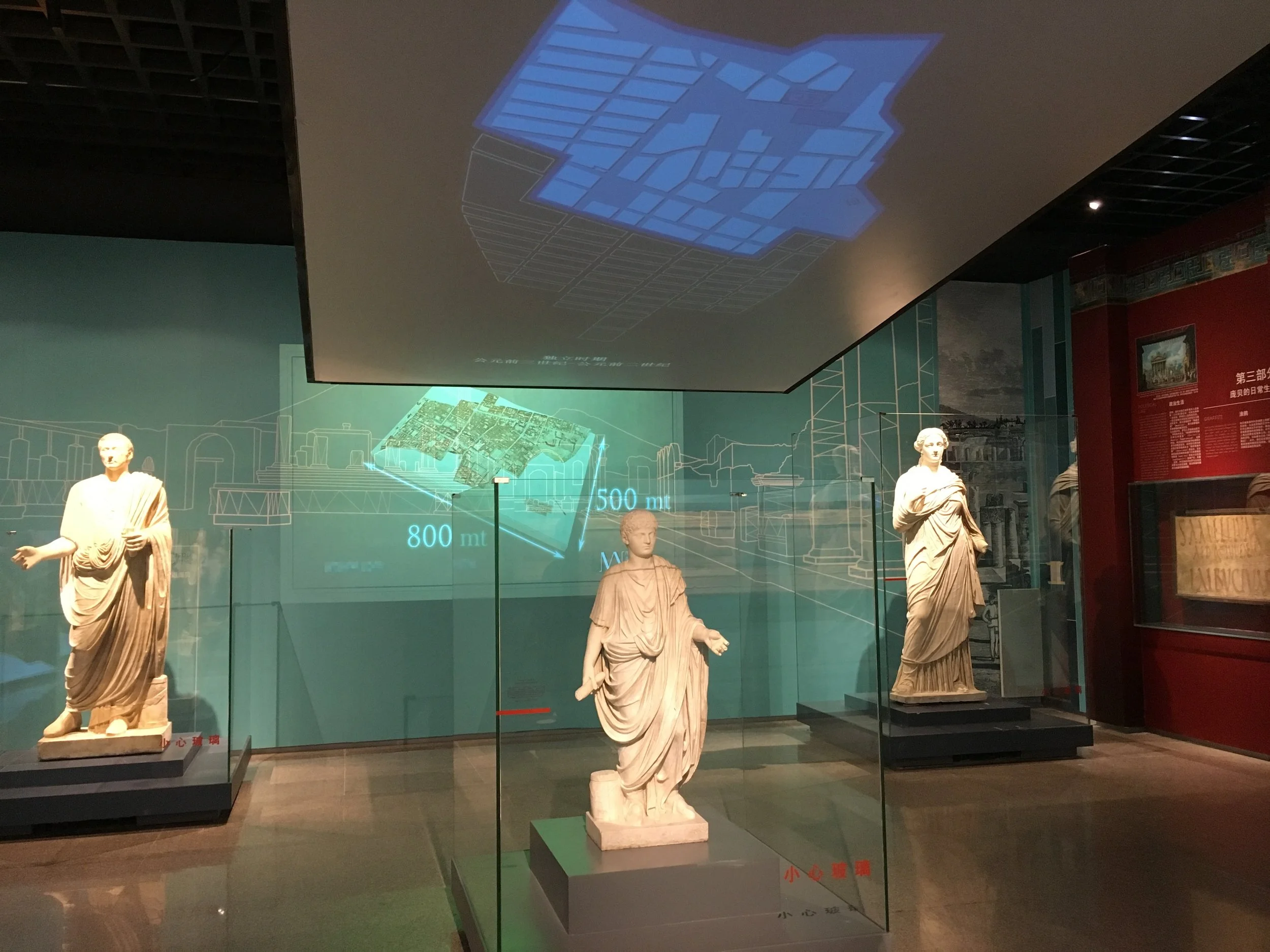 Three ancient Greek statues displayed in a museum, with digital and infographic projections on the background wall. The statues are behind glass cases and are located in a dimly lit room.