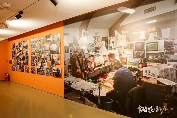 An art gallery wall with a large mural of a person working at a cluttered desk surrounded by artwork and supplies. The mural covers an entire wall, with an adjacent orange wall decorated with numerous photographs.