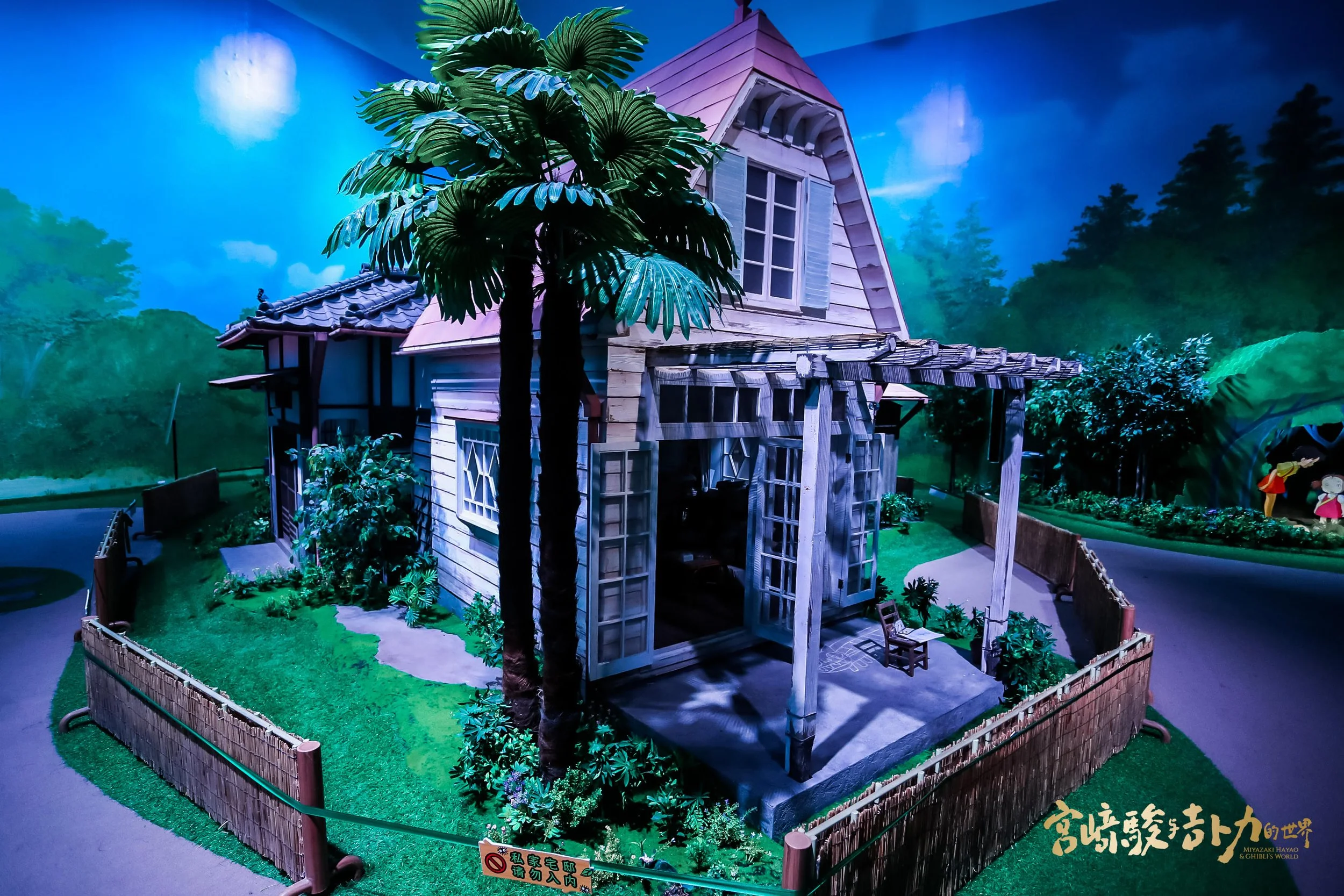 Miniature model of a house with a front porch, surrounded by greenery and flagstone pathway, and a large palm tree in front, set in a landscape with trees and a blue sky background.