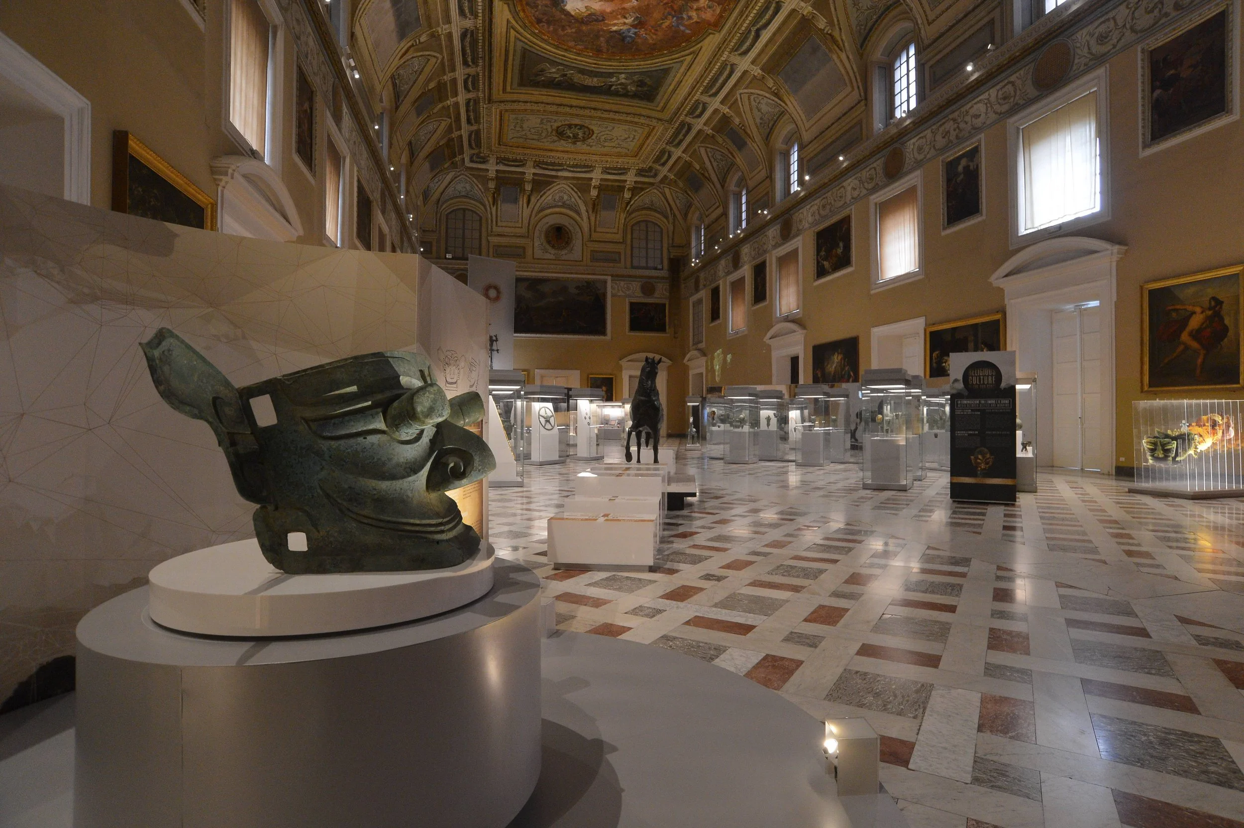 Museum exhibit hall with sculpture of a face in the foreground, glass display cases with artifacts, and a statue of a horse in the background, ornate ceiling, and framed artwork on the walls.