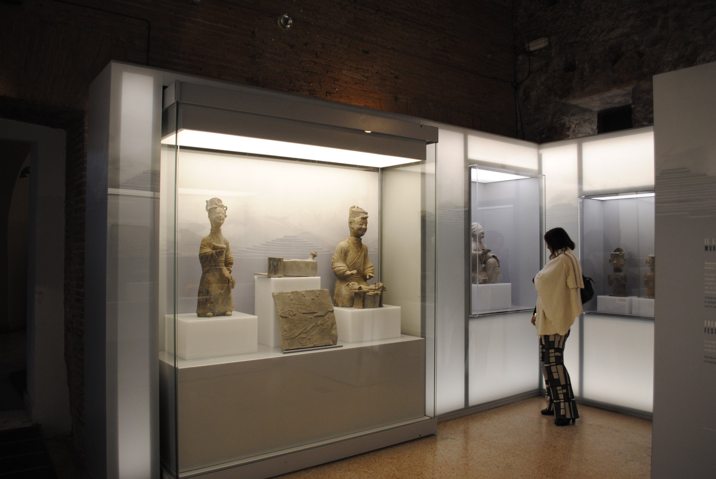 A person in a white coat and patterned pants looking at ancient sculptures inside a museum display case.