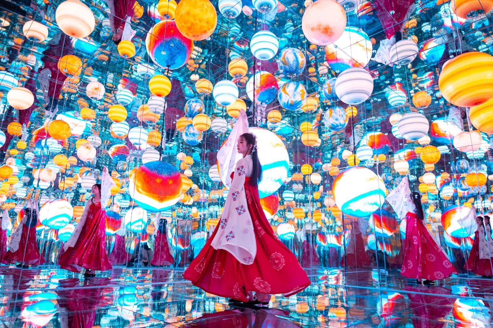 Women in traditional Korean hanbok dresses dancing inside a room filled with hanging and mirrored lanterns of various colors and sizes, creating a vibrant and reflective environment.
