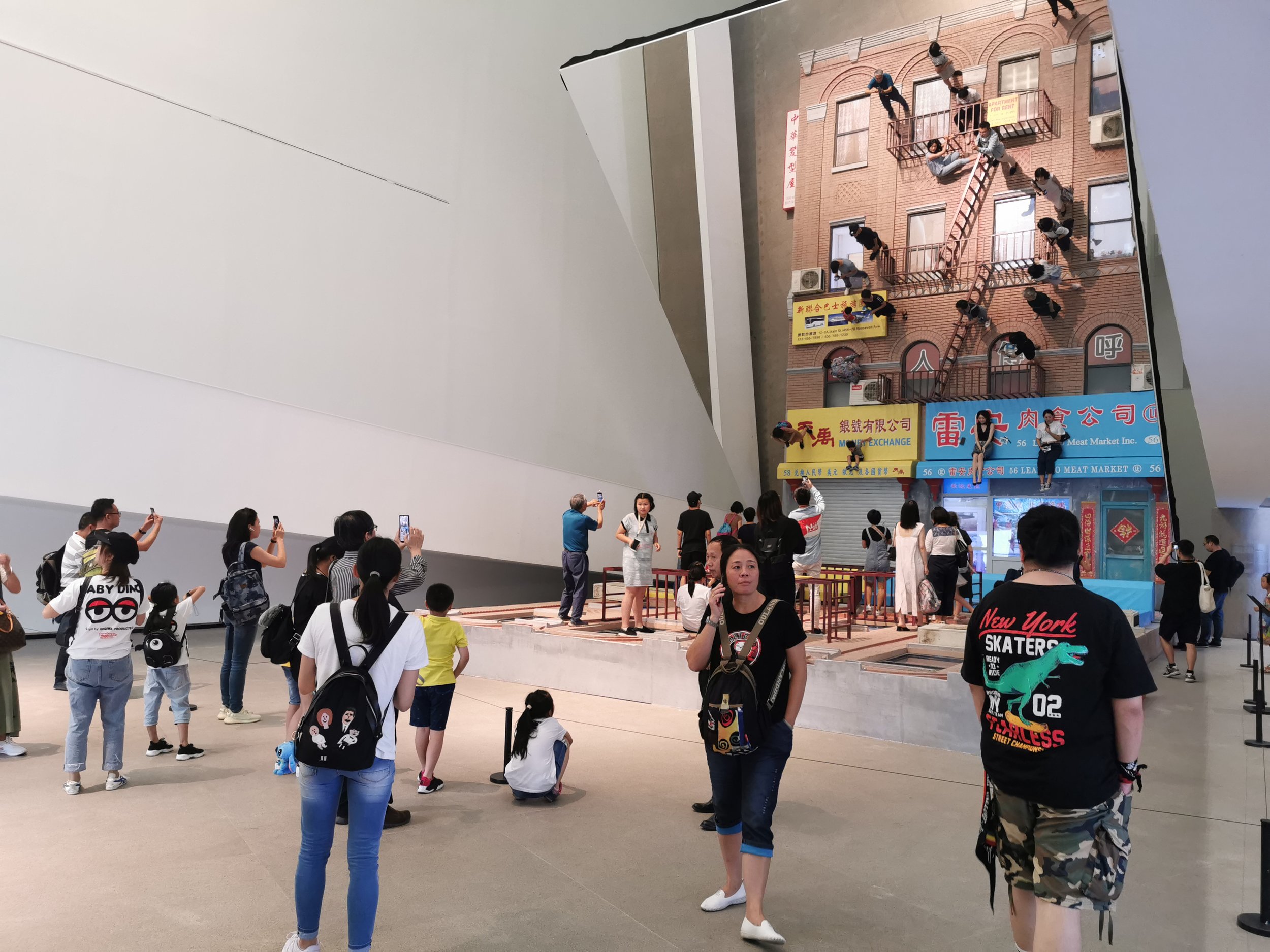People in an indoor space observing a large wall with a photo of an apartment building facade. Some individuals are taking photos with their phones, while others are standing or sitting nearby.