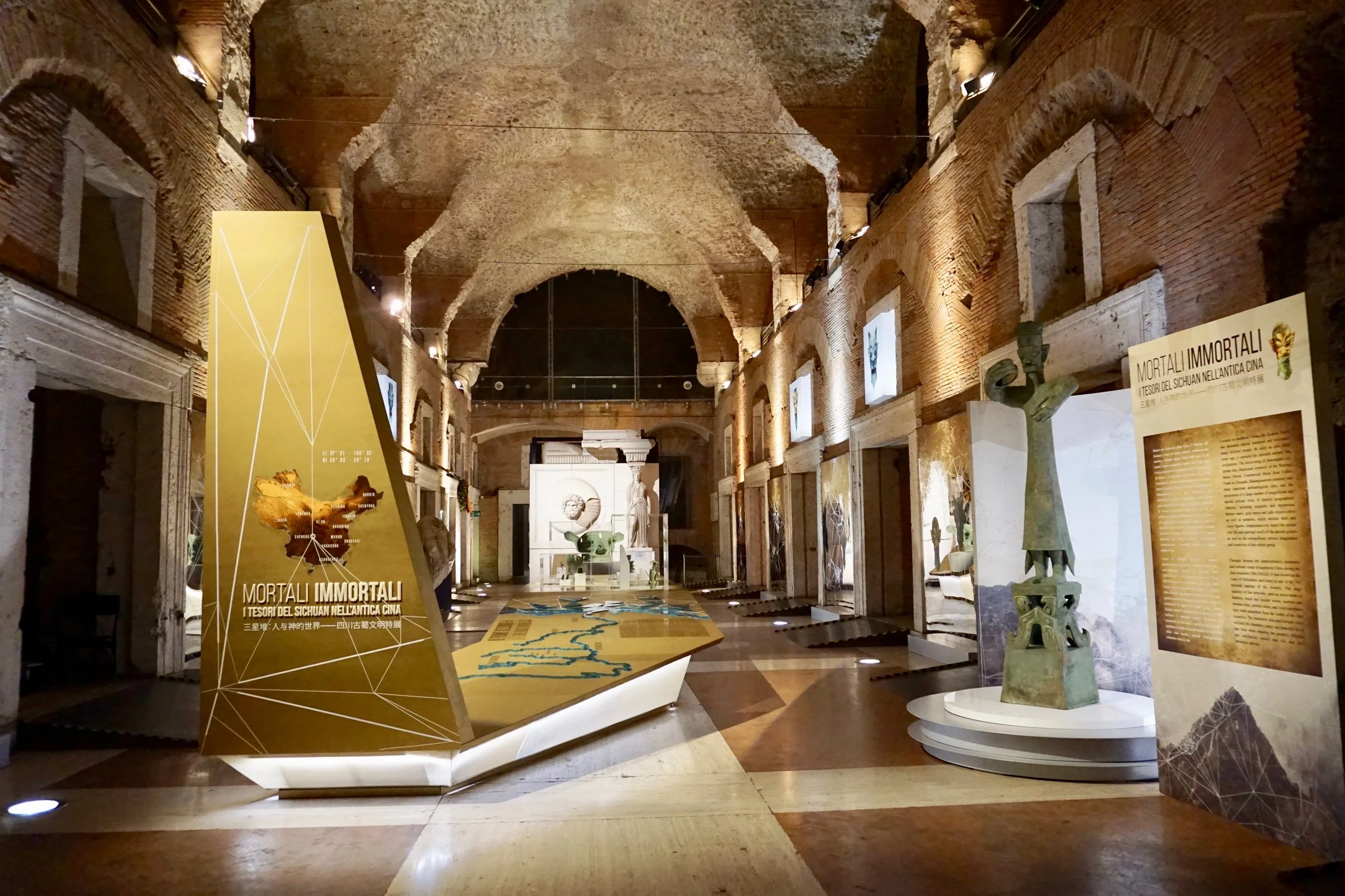 Exhibit hall with ancient sculptures, informational boards, and a large display promoting 'MORTALI IMMORTALI,' featuring a map and text on ancient Chinese civilization, inside a historical building with brick walls and vaulted ceilings.