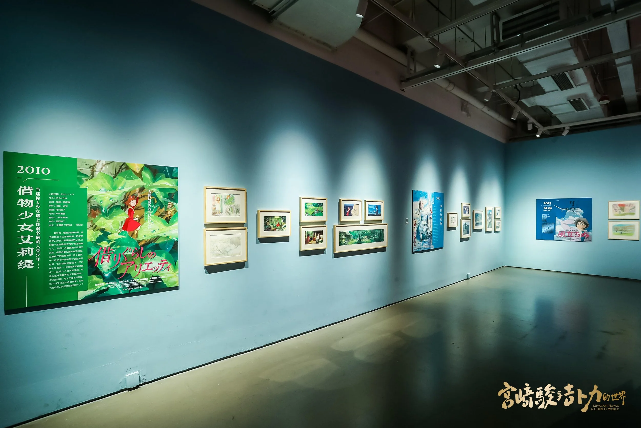 An art gallery exhibition featuring various framed paintings and posters on a light blue wall, with some artwork portraying outdoor scenes and characters. A green poster on the left side includes Japanese text and a colorful illustration of a girl in