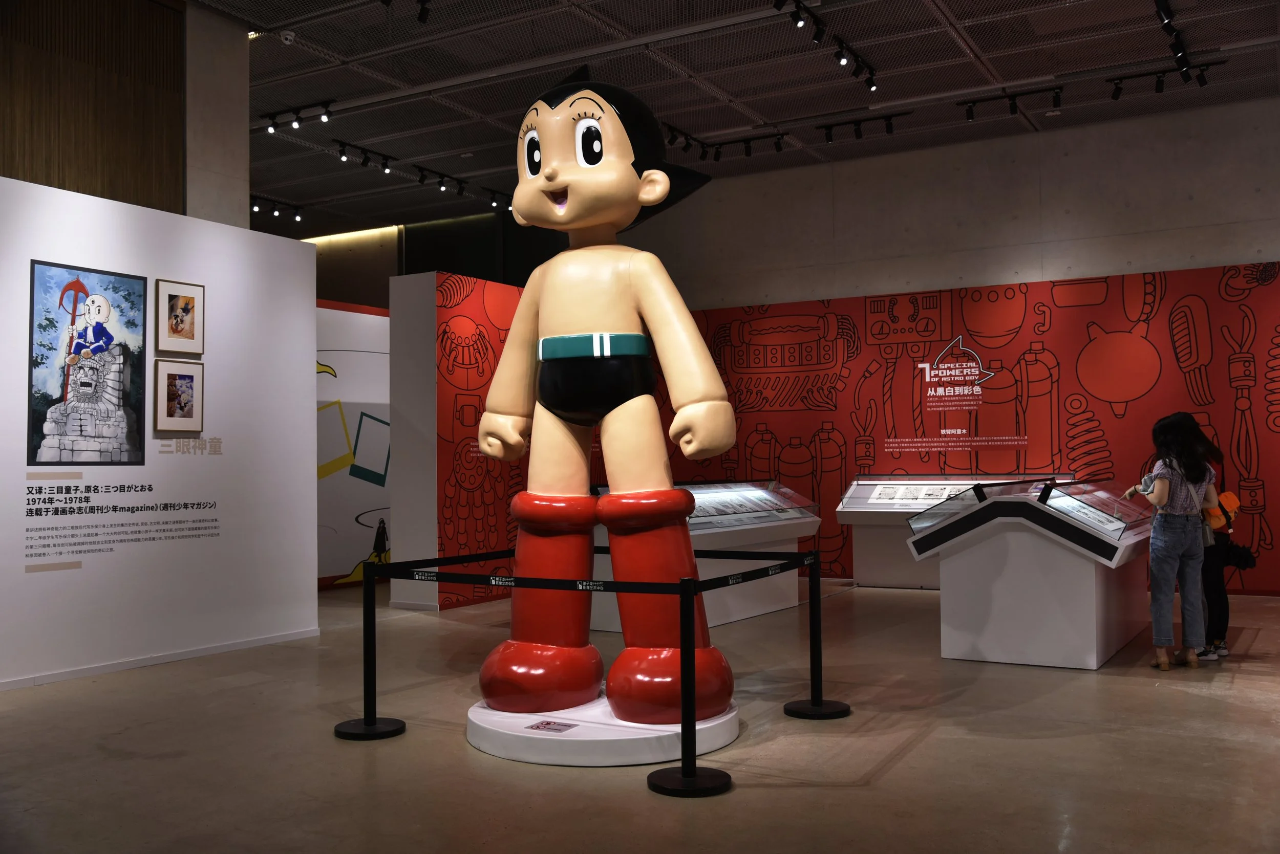 A large cartoonish statue of Astro Boy, a Japanese manga character, displayed in a museum. The sculpture features Astro Boy with a big head, smiling face, black hair, and red boots, wearing black shorts and a green belt. The background includes exhib