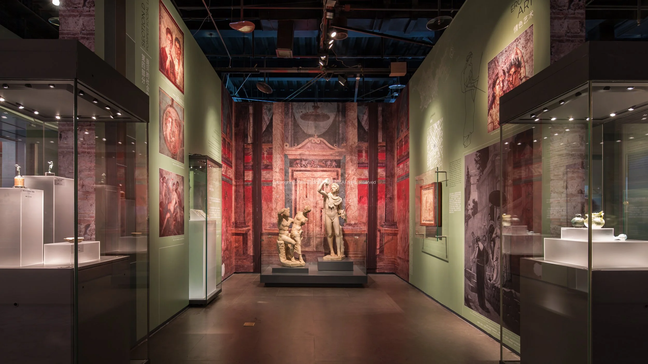 Exhibit of classical sculptures in a museum, including a reclining figure and a standing woman, with wall murals depicting historical artwork and artifacts.