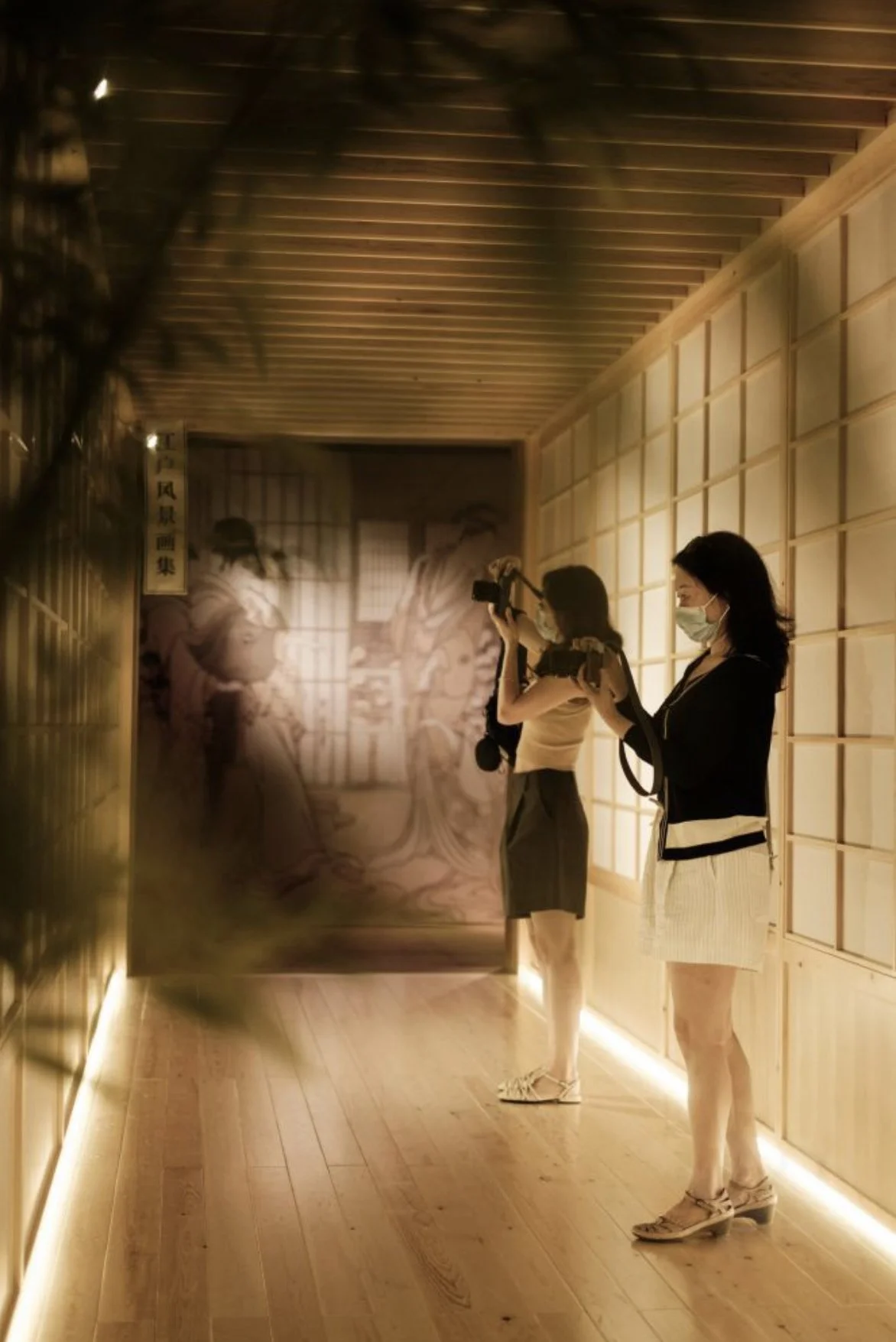 Two women wearing face masks and casual outfits are photographing or filming artwork or exhibition display inside a warmly lit wooden gallery or museum. One woman is holding a camera, and the other a smartphone. The background features a large wooden