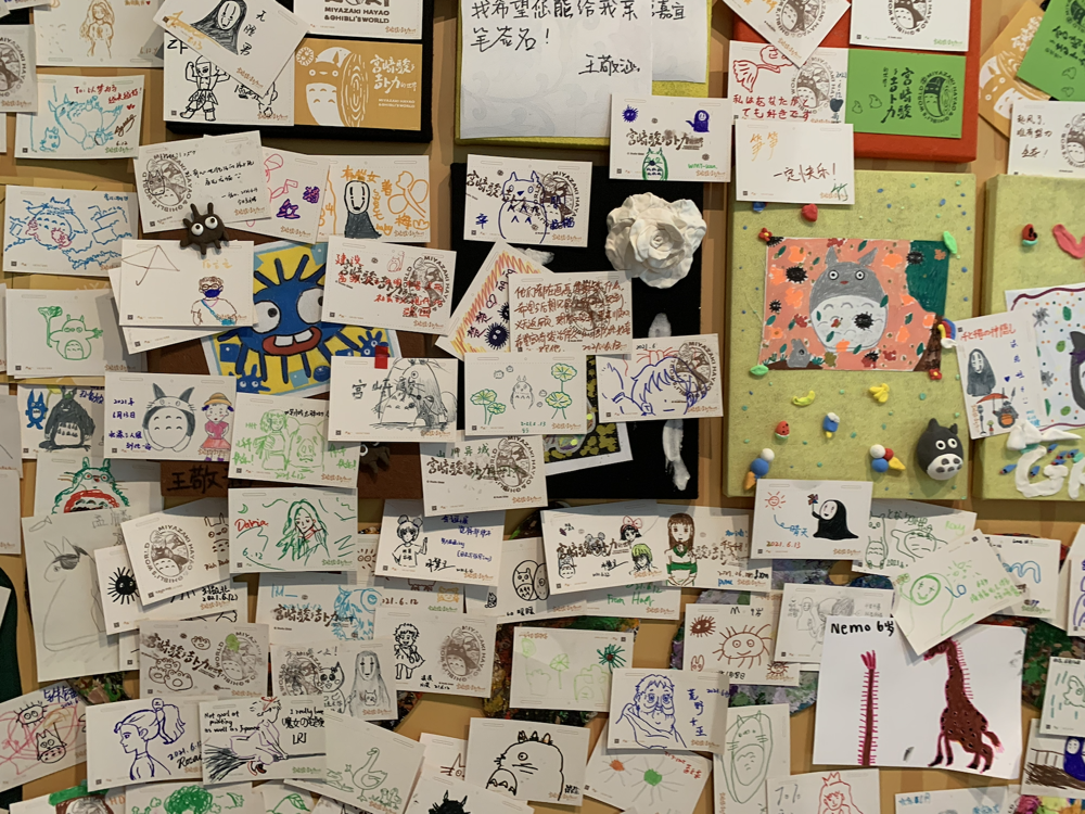 A wall covered with numerous colorful hand-drawn notes, sketches, and small objects, including doodles of animals, characters, and messages, some with stamps, sticky notes, and small decorative items like a toy insect and clay figures.