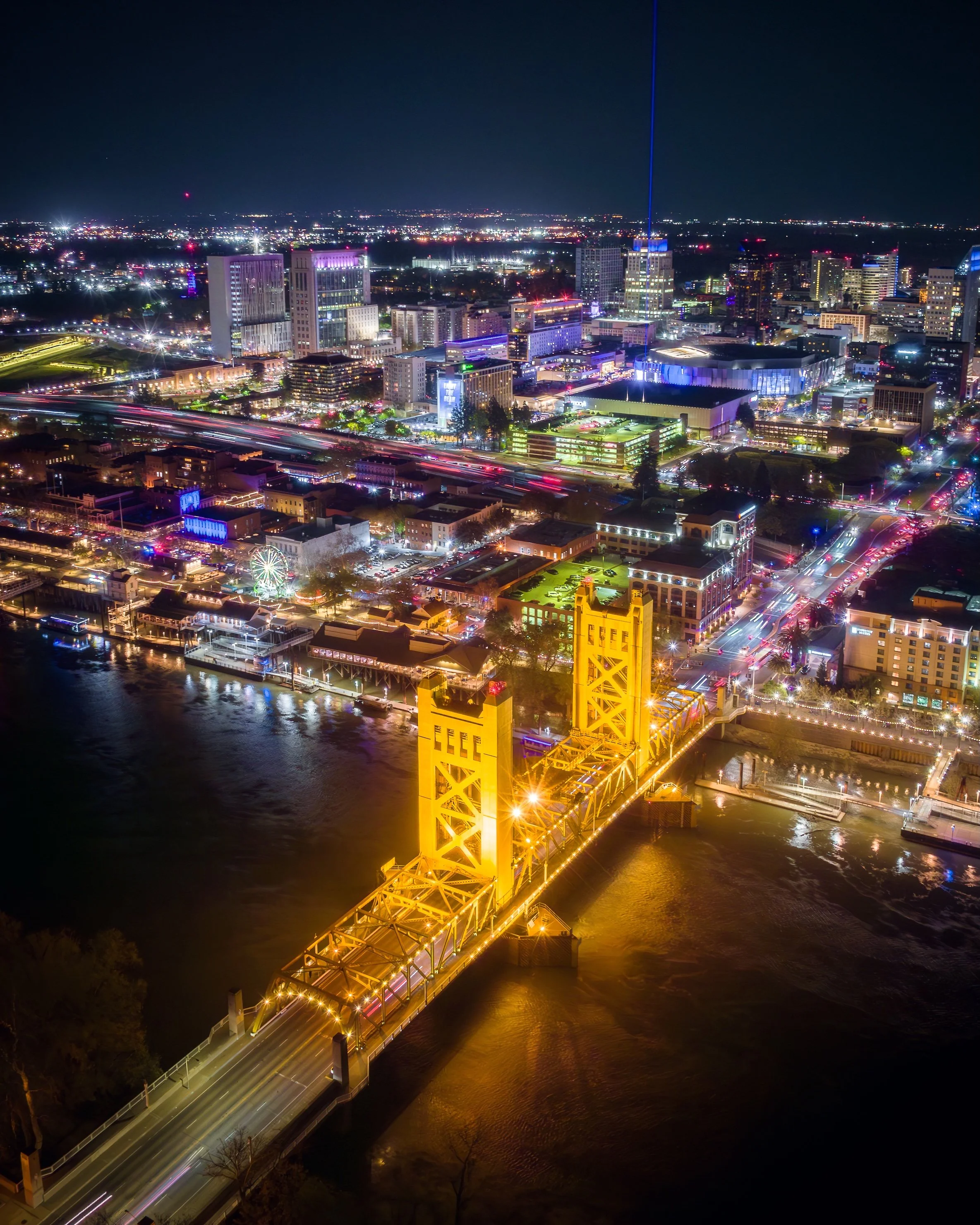 Night view of a city featuring a brightly lit yellow bridge over a river, with a vibrant downtown area filled with colorful lights and tall buildings in the background.