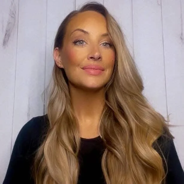 A woman with long, wavy blonde hair and light makeup smiling slightly, wearing a black top and standing against a wooden wall background.