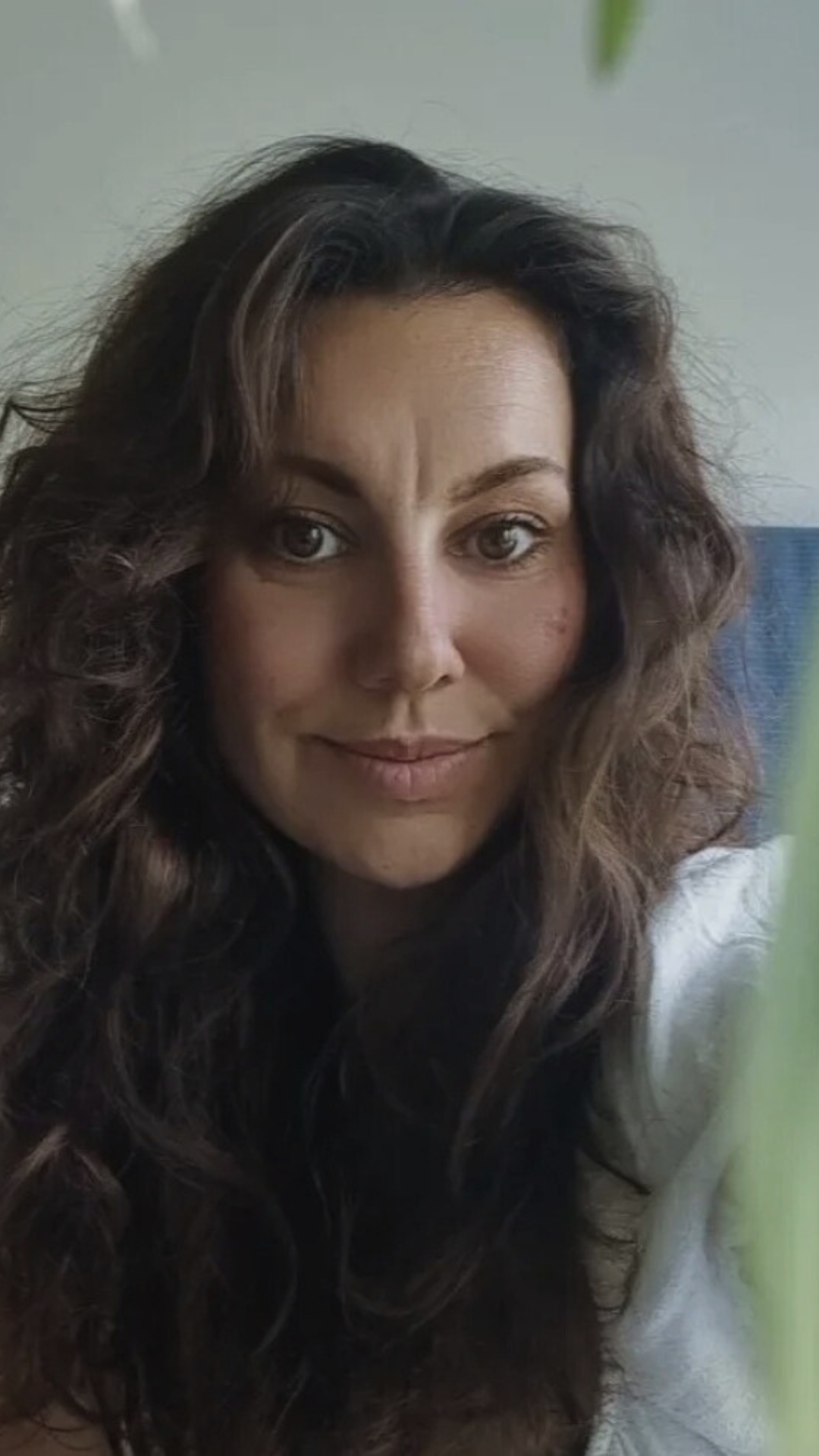 A woman with curly brown hair and light skin smiling, with a blue background and some green leaves visible in the frame.