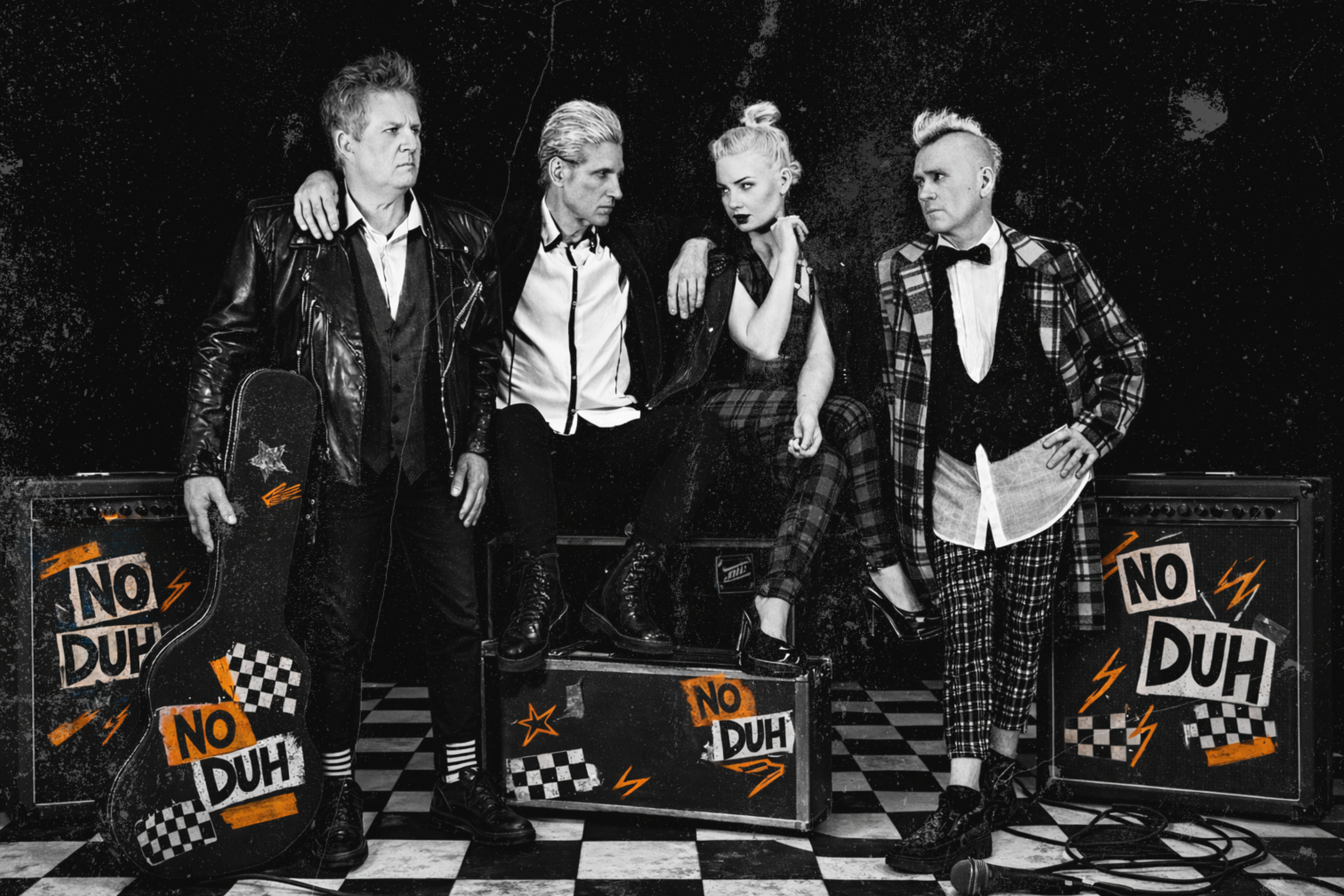 Black and white photo of four individuals standing on a checkered floor behind music equipment. They have punk or alternative fashion styles, with one holding a skateboard. The background has signs with phrases like 'No Duh'.