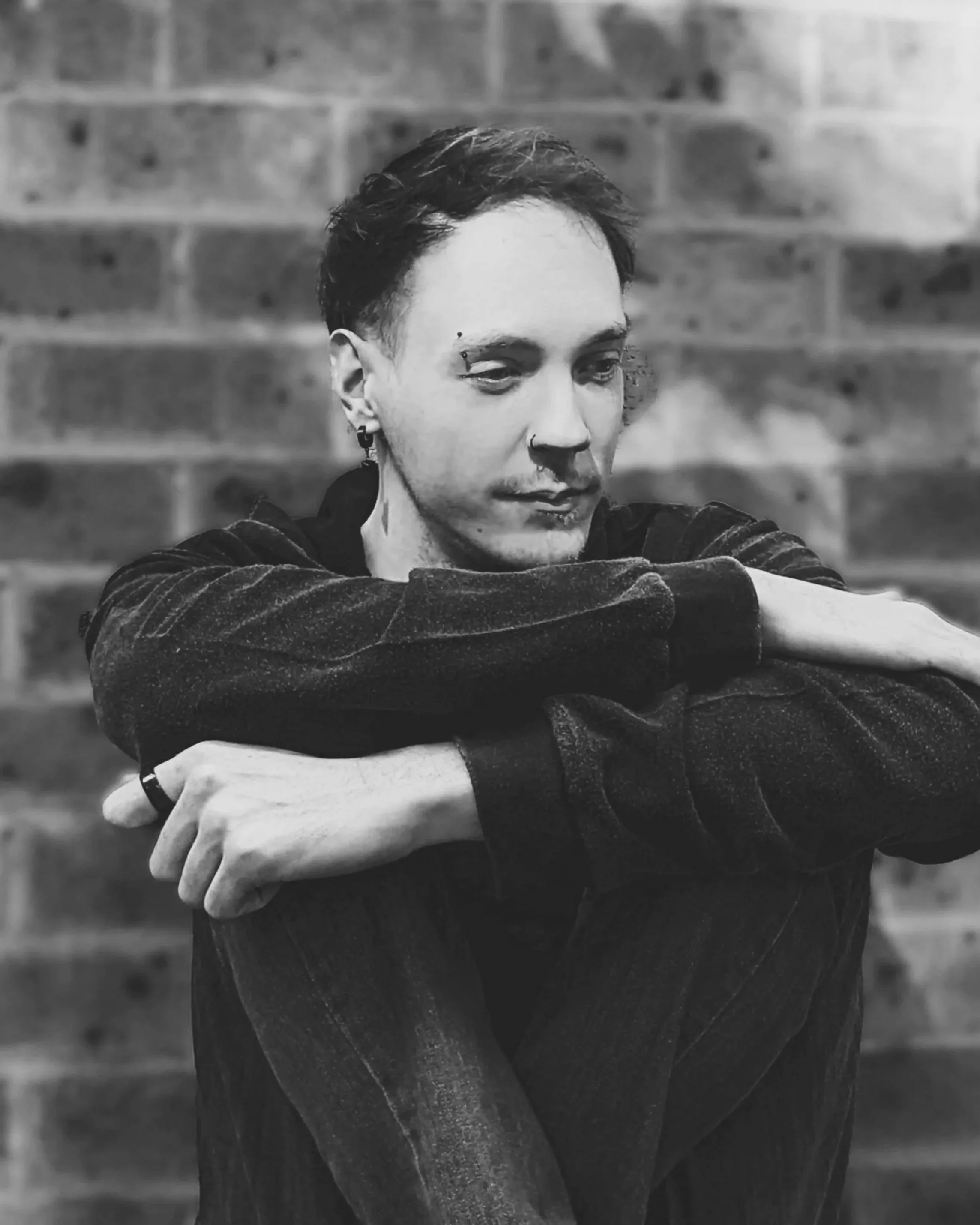A person with short dark hair sitting against a brick wall, wearing dark clothing, with arms resting on knees and looking downward, in black and white.