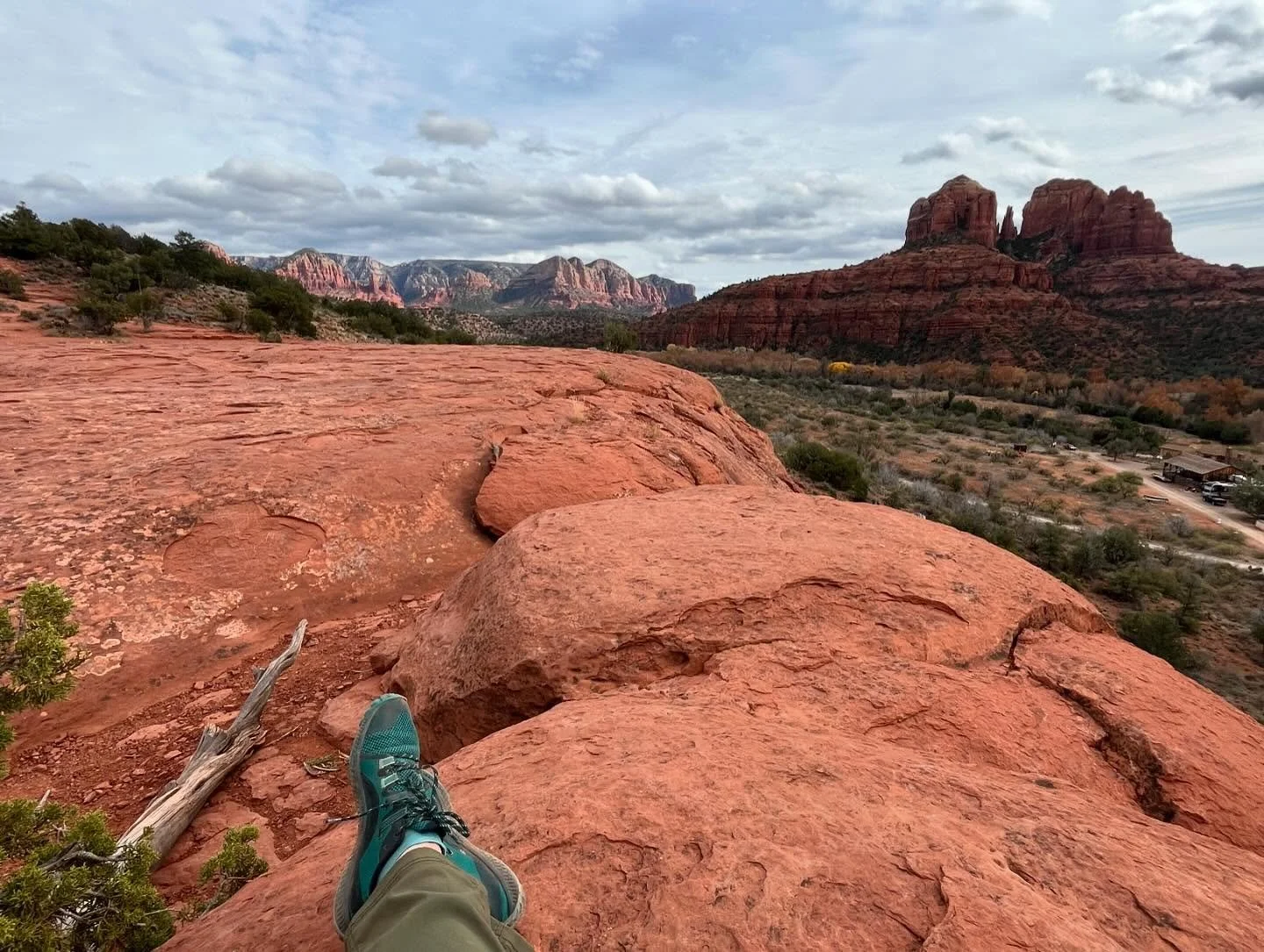 View from a rocky ledge showing a person's legs and blue hiking shoes, with a vast desert landscape, red rocks, green shrubs, and distant mountains under a cloudy sky.