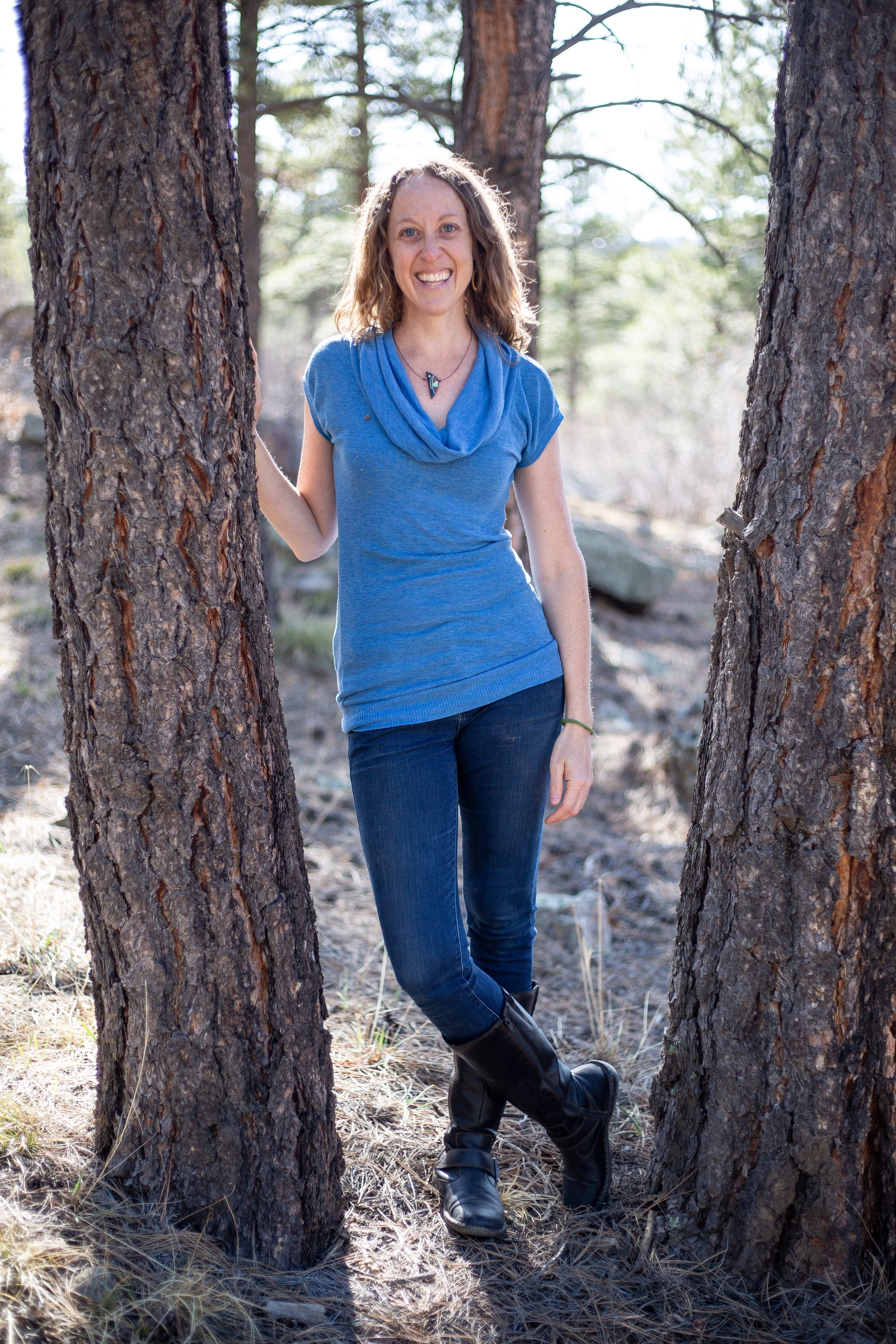 A woman with curly hair, smiling, wearing a blue top, jeans, and black boots, standing outdoors between two trees in a forest, looking at the camera.