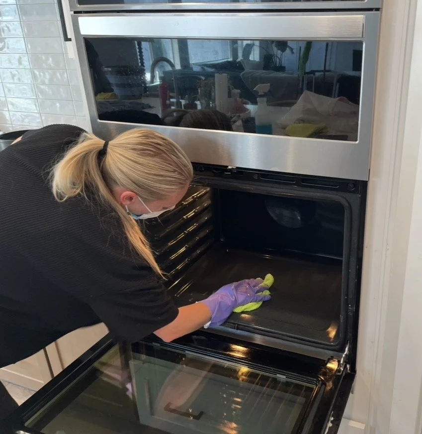 Person wearing a mask and purple gloves cleaning an oven equipped with a microwave above.