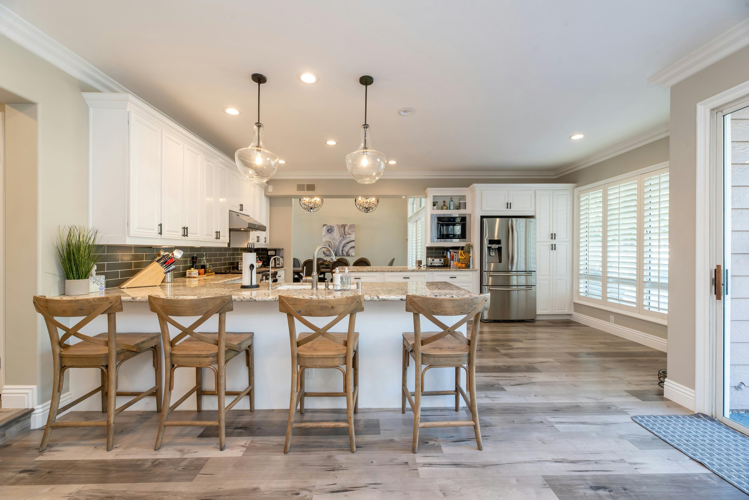 Modern kitchen with white cabinets, stainless steel appliances, a marble island with four wooden chairs, and large windows with plantation shutters.