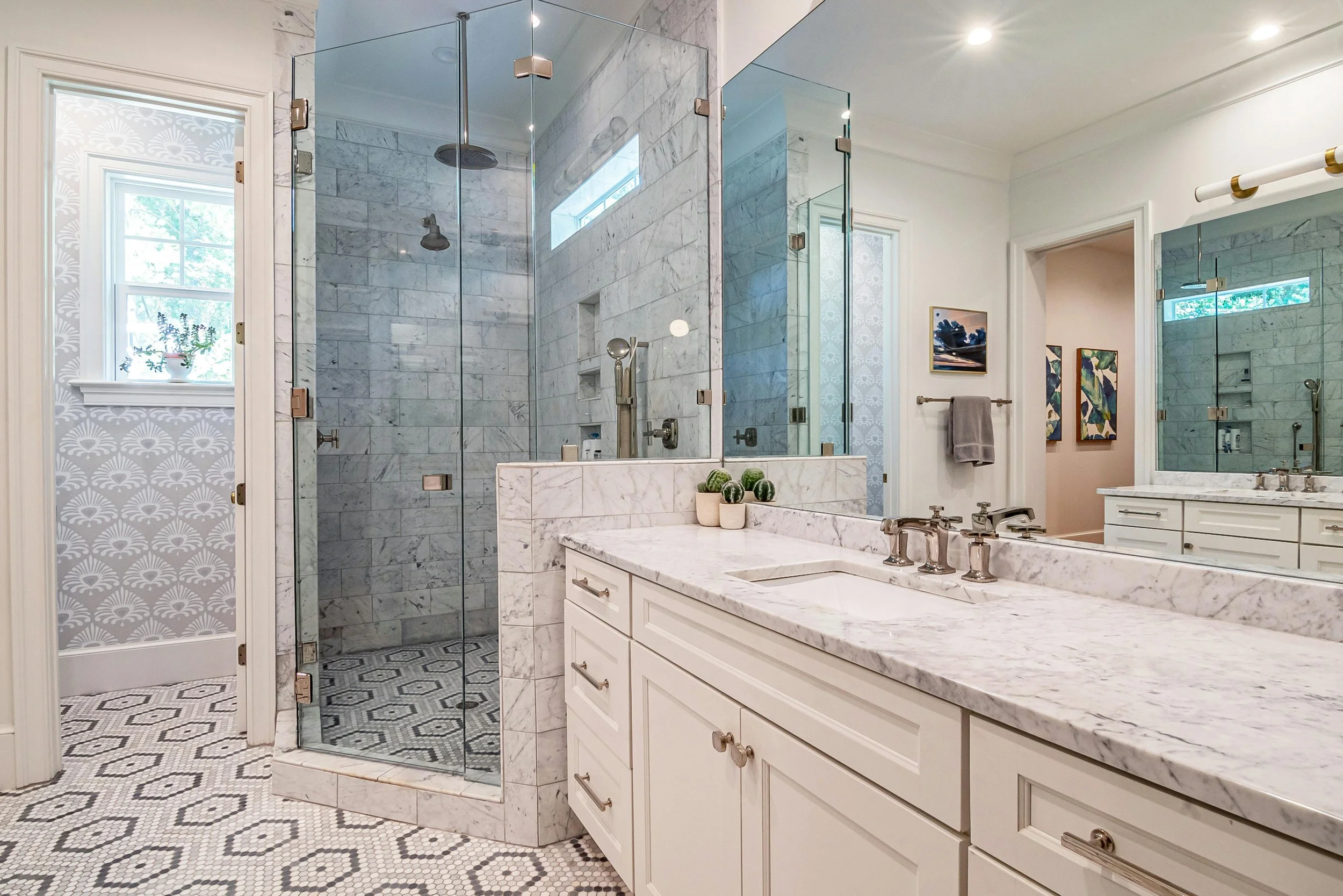A bathroom featuring a marble countertop with a sink, a large mirror, two framed art pieces on the wall, a towel rack with a gray towel, a glass-enclosed shower with marble tiles, a window with a plant, and intricate mosaic tile flooring.