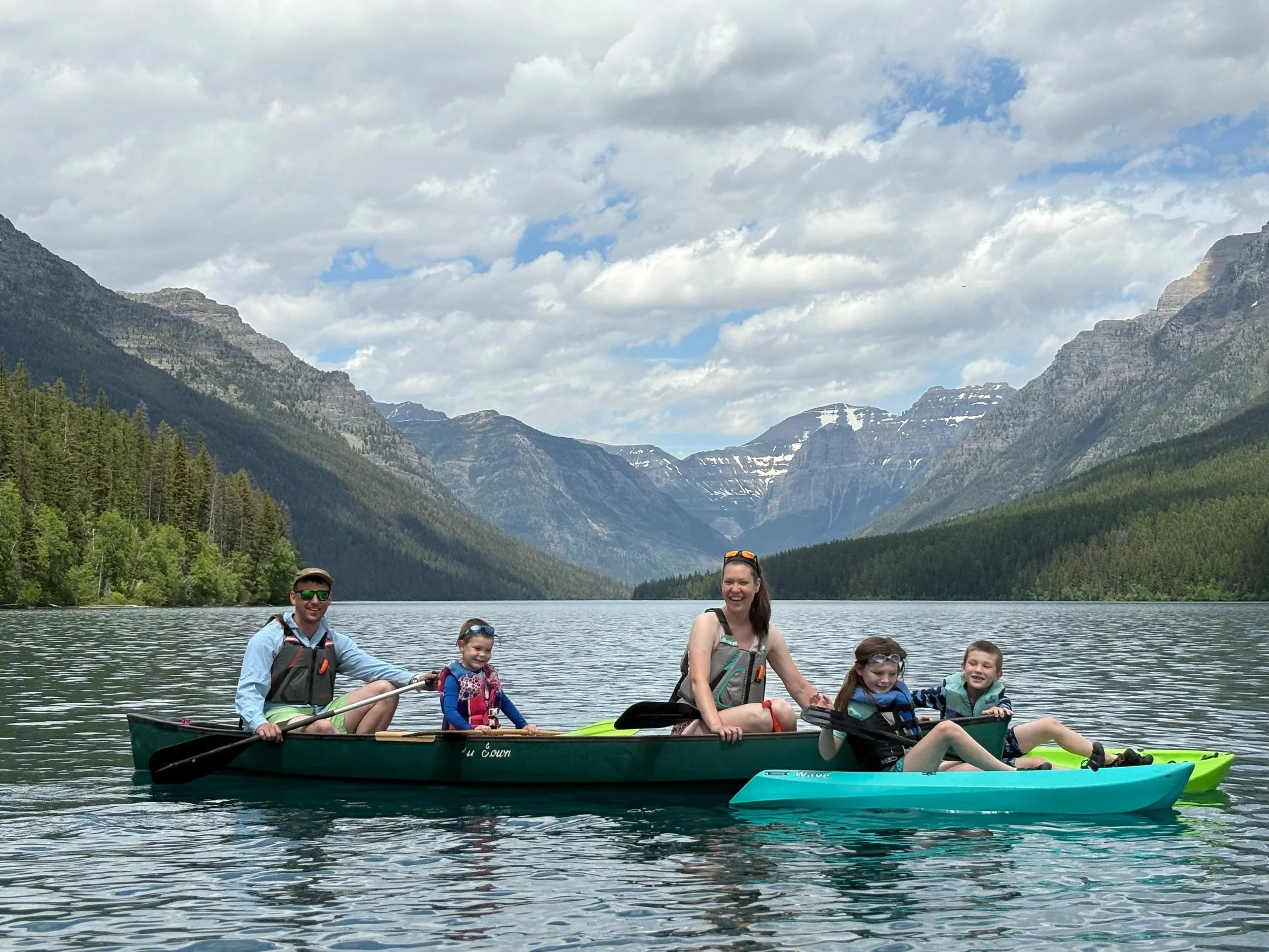 A group of six people, including children and adults, sitting on a canoe and a kayak on a mountain lake with pine trees and snow-capped mountains in the background.
