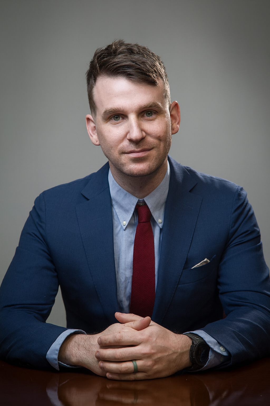A man in a navy suit with a light blue shirt and red tie sitting at a wooden table with hands clasped.
