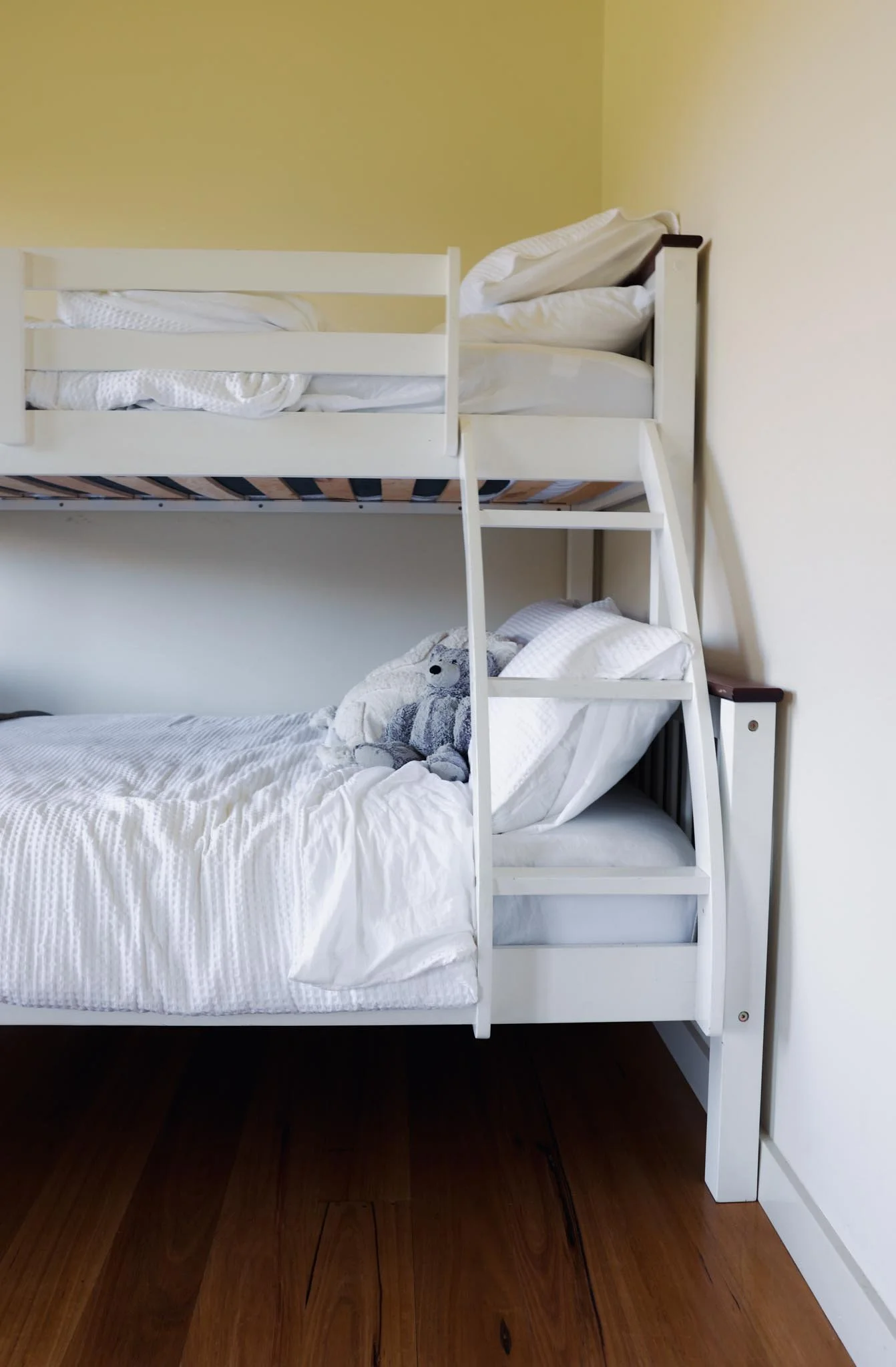 A white bunk bed with a ladder, with the top bunk made and the bottom bunk partially made with a stuffed bear on the pillow. The room has wood flooring and yellow walls.