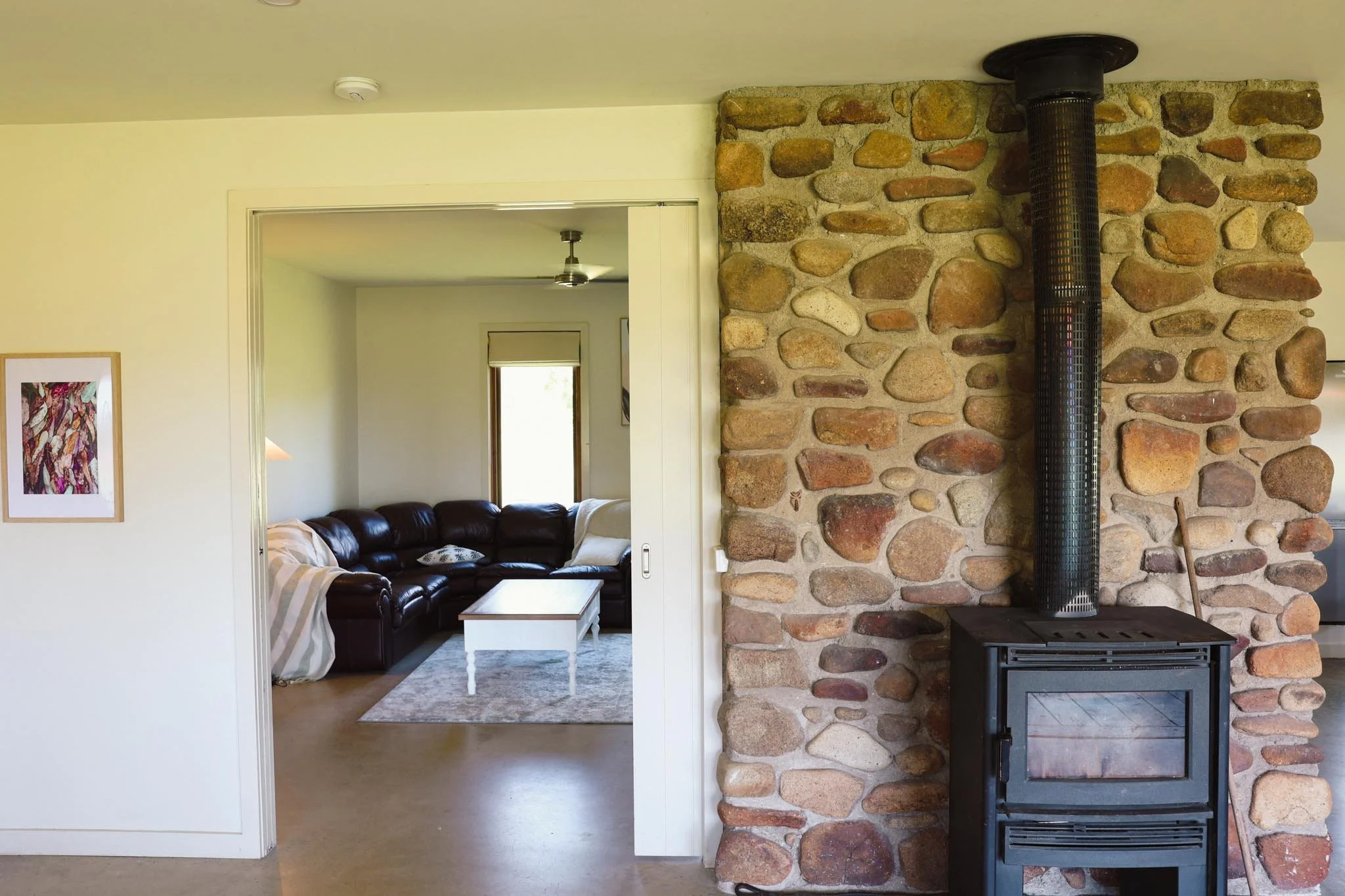 A living room with a stone fireplace and a wood stove, visible through an open doorway leading into a sitting area with a brown leather sectional sofa, a white coffee table, and a window letting in natural light.