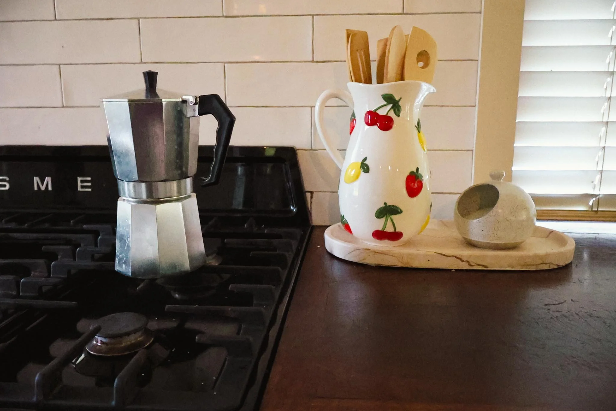 Coffee maker, decorative pitcher with cherries and lemons, and a small stone vessel on a wooden countertop in a kitchen.