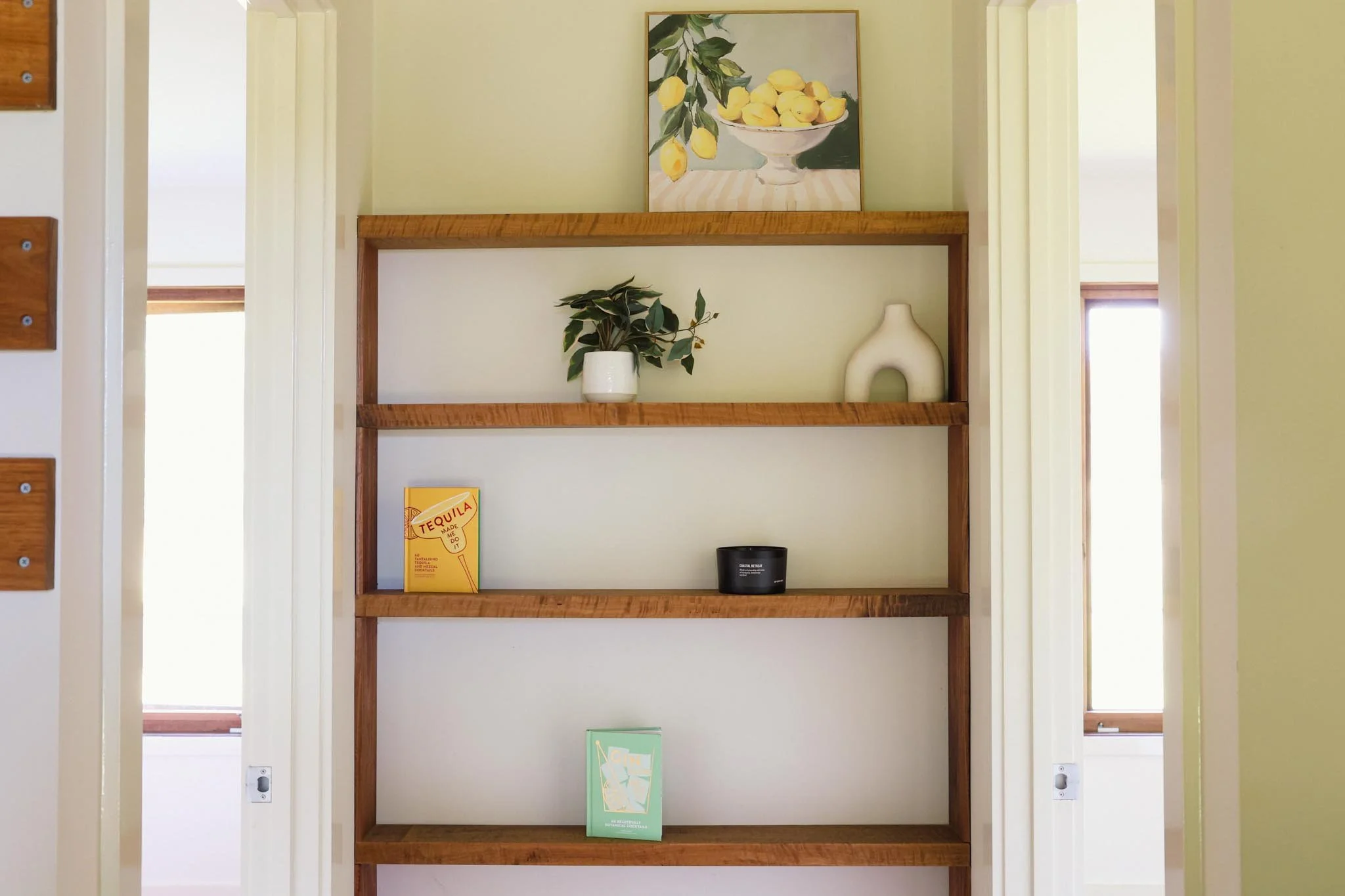 Wooden bookshelf with four shelves, decorated with a fruit painting, potted plant, a white pottery piece, a yellow tequila box, a black candle, and a green book, in a room with cream walls and windows on either side.